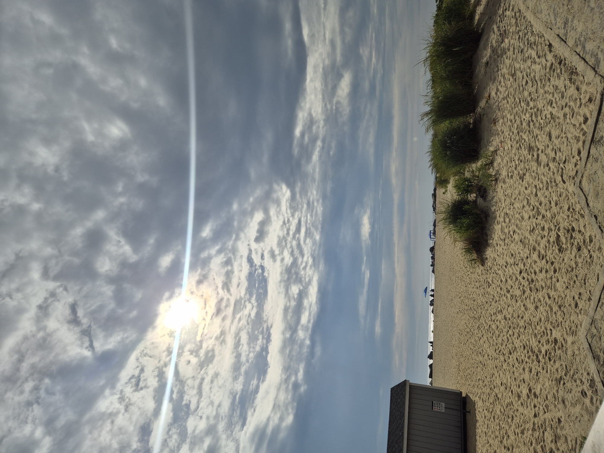 The image depicts a sandy beach scene in Middletown Township, United States of America. In the foreground, on the left, is a small, gray wooden structure with a dark roof. A sign with text, partially obscured, is affixed to the side of the structure. The beach itself is covered in fine, light-brown sand with visible footprints and small undulations. To the right of the structure, a cluster of tall, green grass grows on a sandy dune. In the midground, beyond the beach, the ocean is visible, with a calm surface reflecting the sky. A blue umbrella stands out on the beach near the water's edge, and a few indistinct figures can be seen seated or standing in the distance. The sky above is filled with dramatic, textured clouds, with the sun peeking through, creating a lens flare effect that streaks across the image. The overall atmosphere suggests a possibly overcast but bright day at the beach.