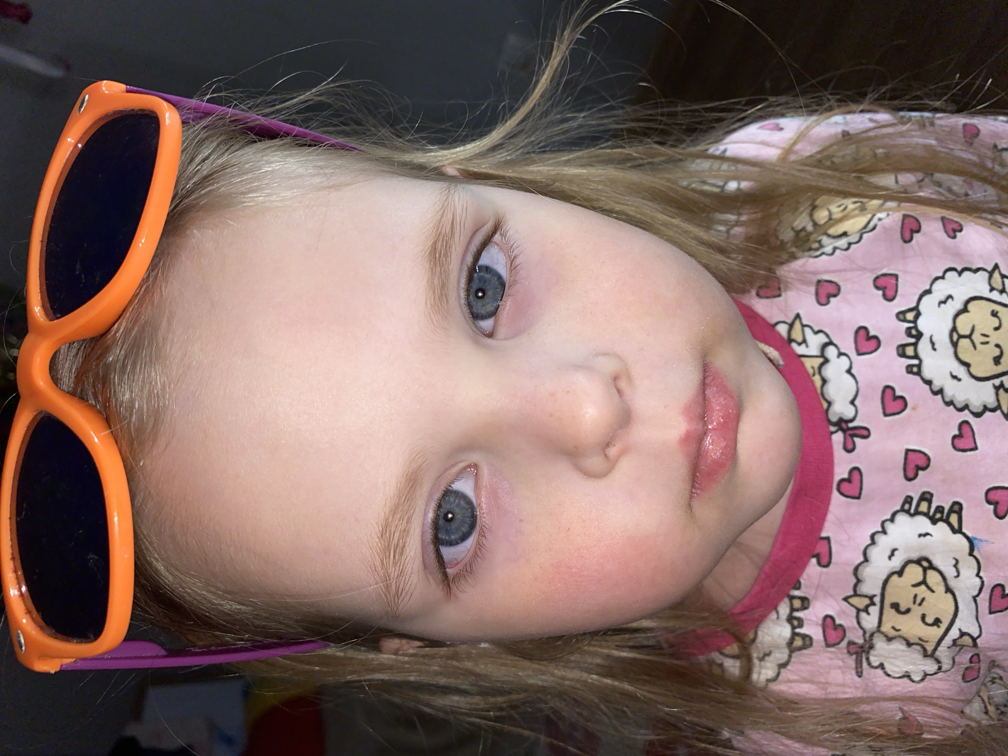 A close-up, slightly tilted portrait shows a young girl with fair skin, blonde hair, and blue eyes. She is wearing orange sunglasses with a purple frame pushed up on her forehead. She has a pink t-shirt with a sheep and heart pattern. The location of Votkinsk, Russia cannot be confirmed from the image.