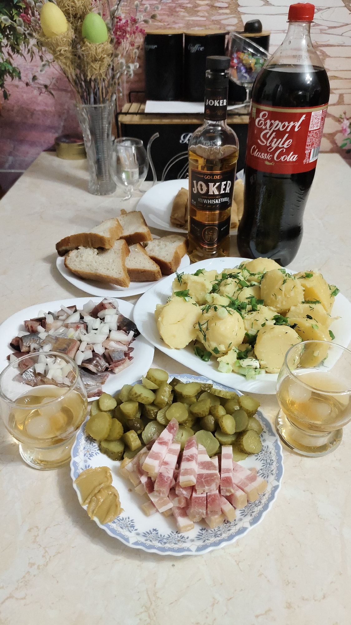 An overhead view captures a table laden with food and drinks, suggesting a casual meal or gathering. The scene is set indoors, likely a home, with a patterned tablecloth on a light-colored surface.
In the foreground, a white plate with a blue decorative rim holds neatly diced pickled cucumbers and cubed pieces of what appears to be cured pork fat (salo) or bacon, alongside a dollop of mustard. Two clear glasses, each containing a light amber liquid and ice cubes, are positioned on either side of this plate. To the left, another white plate features sliced pickled herring generously topped with diced white onions, and behind it, slices of white bread are arranged on a third plate. On the right, a large white plate presents boiled potatoes, cut into chunks and garnished with fresh dill and chopped green onions.
Dominating the center-right is a tall, dark brown bottle of "GOLDEN JOKER" labeled "with WHISKEY." Next to it stands a large red-labeled bottle of "Export Style Classic Cola." In the background, decorative elements include a glass vase filled with dried branches, some adorned with small yellow and green egg-shaped ornaments, possibly hinting at an Easter theme. Further back, two black canisters with white labels "Sugar" and "Coffee" are visible on a metal rack, along with a small clear glass creamer.
No people are visible in the image. The arrangement of food and drink suggests a convivial atmosphere, possibly an informal celebratory meal or a typical Russian spread, consistent with the Krasnoyarsk, Russia context. Visible text includes "JOKER," "GOLDEN JOKER with WHISKEY," "Export Style Classic Cola," "Sugar," "Coffee," and a small date/time stamp "02.03.24 21:18" on the cola bottle.