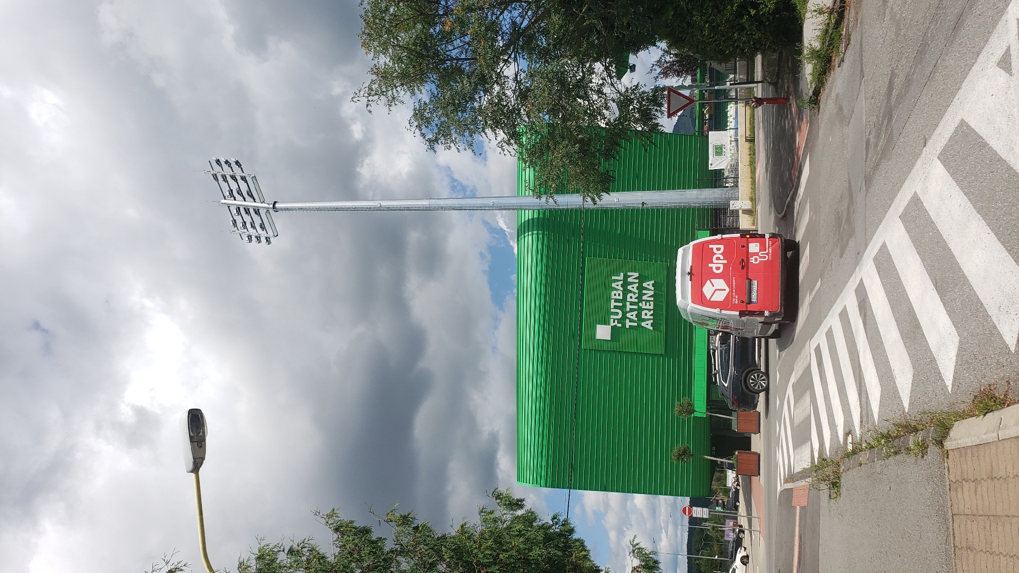 The image depicts the Futbal Tatran Aréna in Prešov, Slovakia, on a cloudy day. A DPD delivery van is seen driving away from the camera on a road that has a pedestrian crossing marked with white stripes. The van is red and white with the DPD logo on the back. To the left of the van, a black SUV is parked. The Futbal Tatran Aréna is a large, green, modern building with horizontal corrugated siding, and the words "FUTBAL TATRAN ARÉNA" are visible on its facade. A tall light pole with multiple stadium lights stands to the right of the arena. In the foreground, there is a sidewalk and some grass growing along the edge of the road. A yield sign is visible in the background to the right.