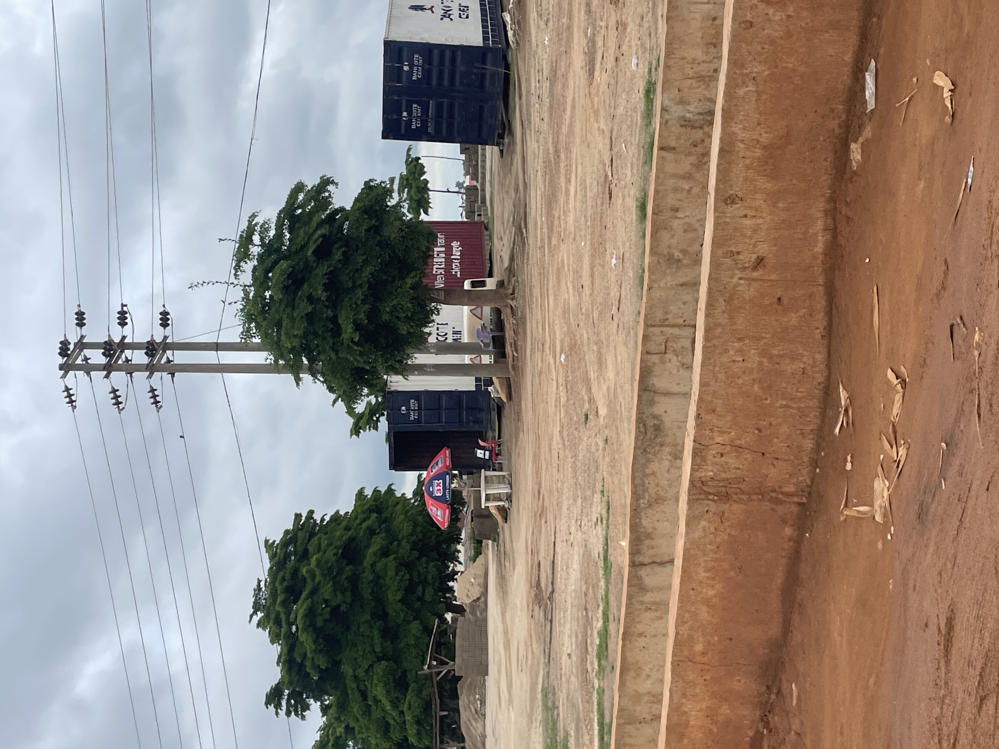 The image depicts an outdoor scene in Minna, Nigeria. In the foreground, there is a reddish-brown dirt ground with some scattered debris. Two concrete barriers run horizontally across the frame, with the closer one being more prominent and showing some signs of weathering and cracking. In the mid-ground, a dusty, open area features several large shipping containers, some dark blue and white, others red. One blue and white container has visible branding that reads "BANJOTE CEM HWT." A red umbrella is open over a white circular table with two white chairs, suggesting a small informal seating area. A tall, grey utility pole with multiple crossbars and insulators supporting power lines stands prominently on the right side of the frame, extending into the overcast sky. Lush green trees are visible, with one large tree in the center-left and another to its right, its branches reaching towards the utility pole. The sky is cloudy and grey. In the far background, there are more trees and some structures, possibly a small building with a thatched roof, and stacks of what appear to be construction materials like bricks. The overall impression is one of an industrial or construction site in an urban or semi-urban environment under an overcast sky.