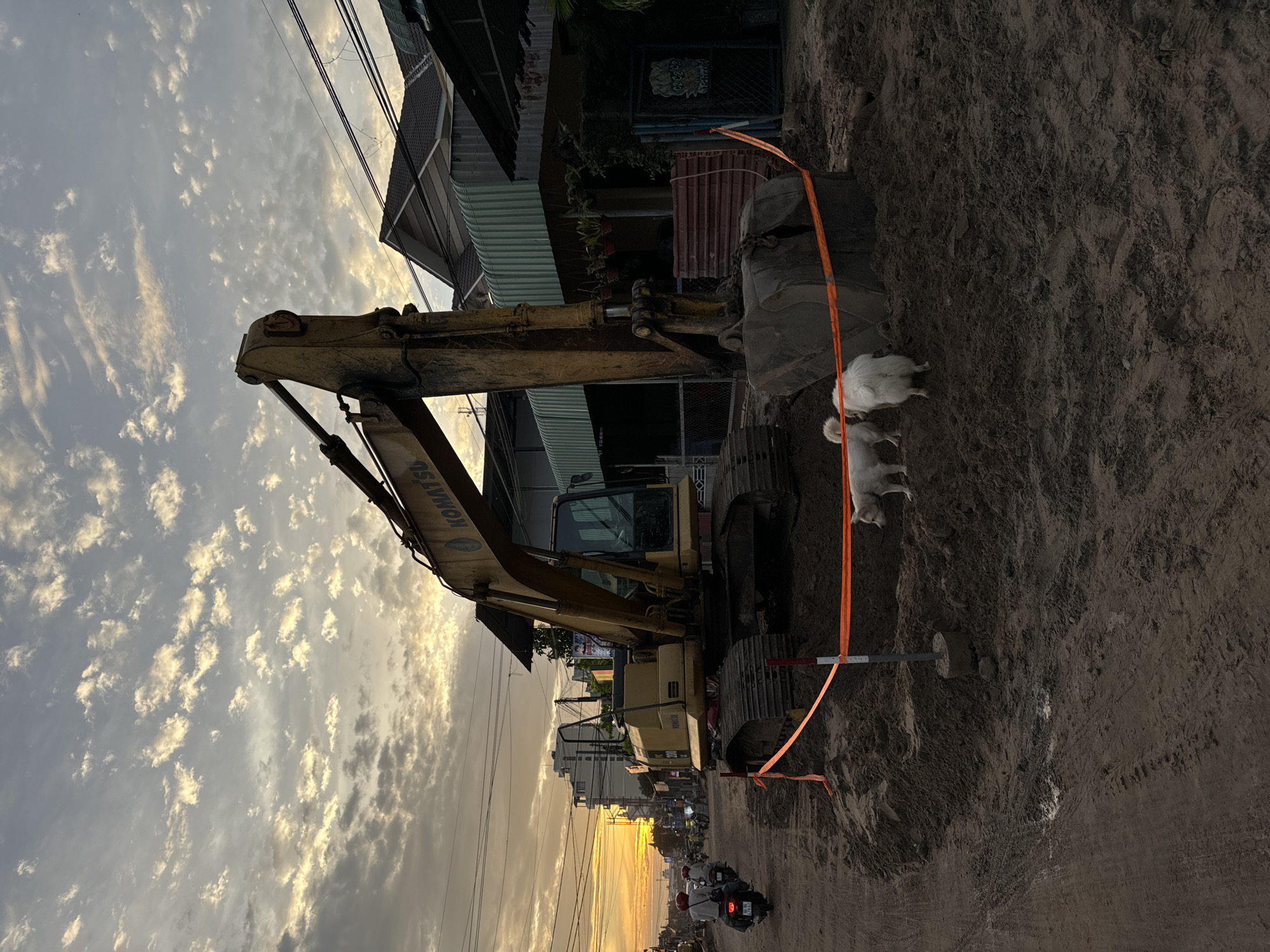 At dusk in Mũi Né, Vietnam, an excavator, identified by the "KOMATSU" logo, is parked on a dirt road, indicating ongoing construction. The sky is filled with scattered clouds, illuminated by the setting sun. Two white dogs are visible near the excavator, one facing away from the camera and the other looking towards the left. The area is cordoned off with orange safety tape. In the background, houses with corrugated metal roofs and power lines are visible, along with several motorbikes on the road. The overall scene depicts a typical street in a developing area during twilight.