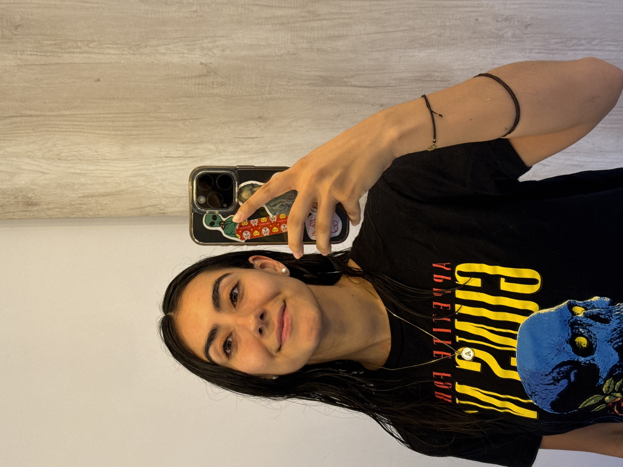 A woman is taking a selfie indoors. She has dark hair and is wearing a black t-shirt with yellow and red text and a blue skull graphic. She is holding a phone with a sticker-covered case. She has a bracelet on her arm and is wearing a necklace. The background includes a white wall and a wood-patterned surface. The location is not specified as Puebla, Mexico in the image.