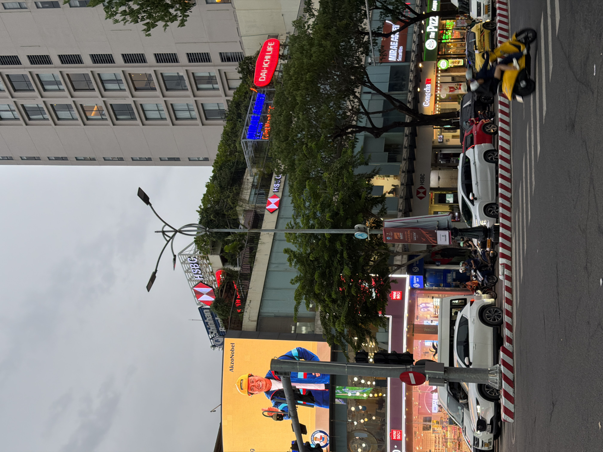 A street scene in Đà Nẵng, Vietnam, features a busy intersection with multiple businesses and traffic. A large digital billboard displays an advertisement for AkzoNobel, featuring a smiling man in a yellow hard hat and blue jacket holding a tool. Above the billboard, a Shinhan Bank sign is visible. Further down the street, a tall, modern building with many windows dominates the skyline, with a large HSBC sign on its lower floors. Trees line the street, and a traffic light is mounted on a pole. Various vehicles, including cars and motorcycles, are on the road, with a yellow scooter and its rider prominently in the foreground. Several commercial establishments are present, including GongCha, The Pizza, and Mirage Asset Securities. The sky is overcast.