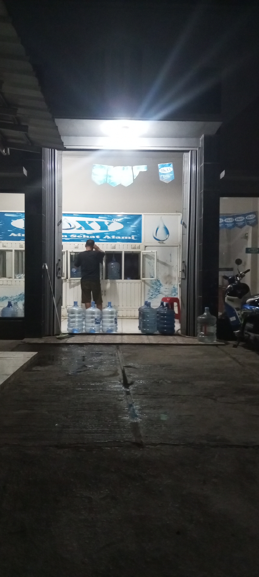 A man stands in front of a store entrance, which is open. The man is wearing a dark t-shirt and shorts. He is facing away from the camera and appears to be adjusting something on his head. Inside the store, there are several large plastic water bottles stacked on shelves. Outside the store, more water bottles are placed on the ground. The store appears to be a water refill station. A motorcycle is parked to the right of the entrance. The scene is dimly lit by overhead lights. The store has a blue and white sign that says "AIR MINUM SEHAT ALAMI," which translates to "Natural Healthy Drinking Water." The location is identified as Setu, Indonesia.