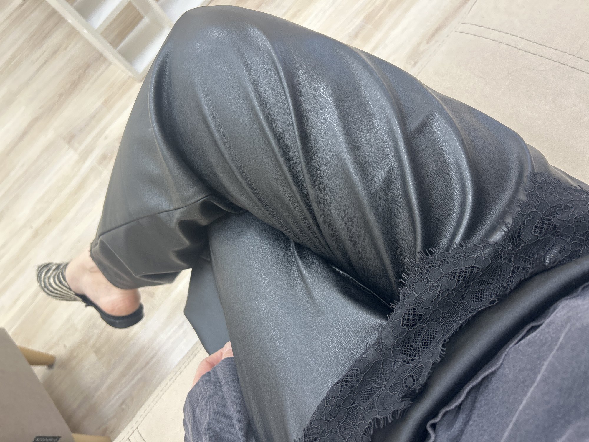 A photograph taken from above shows a person sitting with their legs crossed. The person is wearing black, faux-leather pants and a black top with lace trim. A zebra-striped mule is visible on the foot of the lower leg. The person is sitting on a beige sofa. The floor is light wood-tone. A white bookcase is visible in the background. A chair can be seen in the lower left corner. The location is not specified, but the user asked it be set in the city of Izhevsk, Russia.