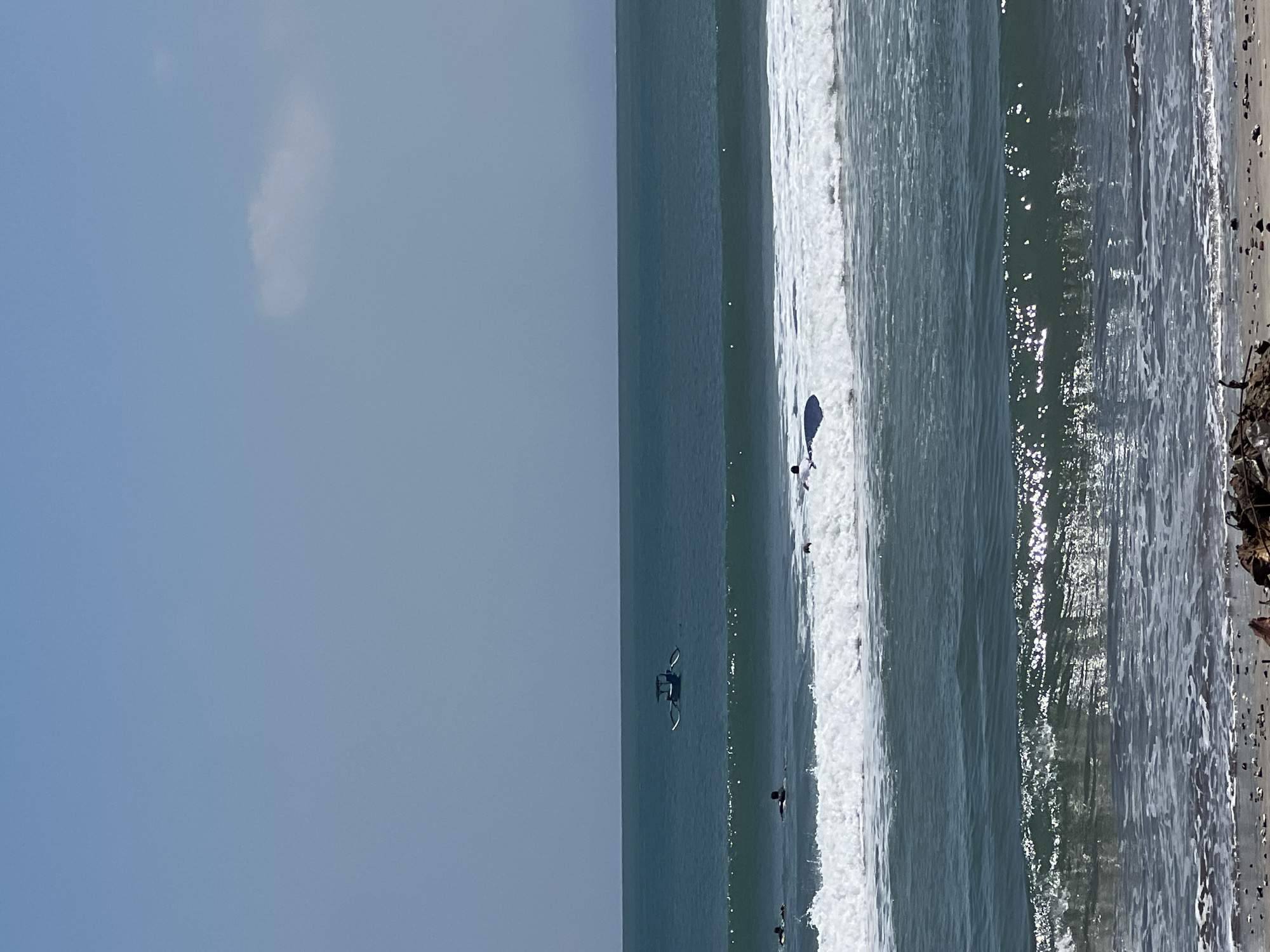 The image depicts a wide shot of the ocean in Kuta, Indonesia, on a bright, sunny day with a clear blue sky. In the foreground, waves are crashing on the shore, revealing a beach covered in small stones and debris. Further out in the water, several surfers are visible, some on their boards and others paddling. A traditional Indonesian outrigger boat, known as a jukung, is anchored in the distance. The water is a deep teal color, with distinct lines of breaking waves creating white foam. A single, wispy cloud floats in the upper right corner of the sky. The scene captures a typical day at the beach in Kuta, known for its surfing spots.