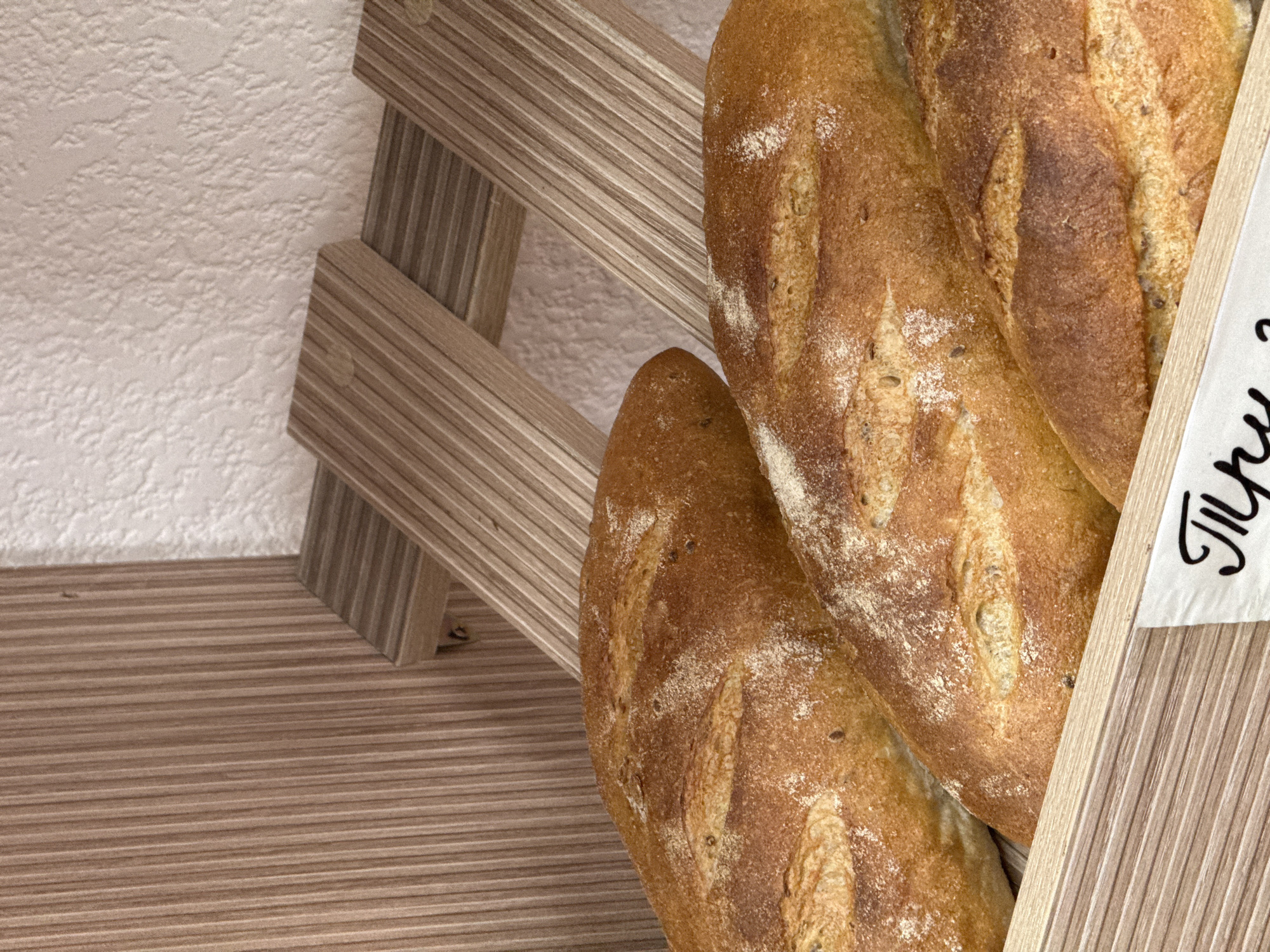 A close-up, eye-level shot showcases three loaves of golden-brown bread in a wooden display crate. The bread has a slightly floured crust with visible scoring marks and small seeds embedded in the surface. The loaves are arranged diagonally in the crate, with the closest loaf filling the bottom right of the frame. The background consists of a ribbed wooden panel on the left and a textured, light-colored wall on the right, with wooden shelves supporting the bread display. A white paper tag with stylized black lettering is visible in the bottom right corner, but the text is partially obscured. The scene suggests a bakery or food market setting.