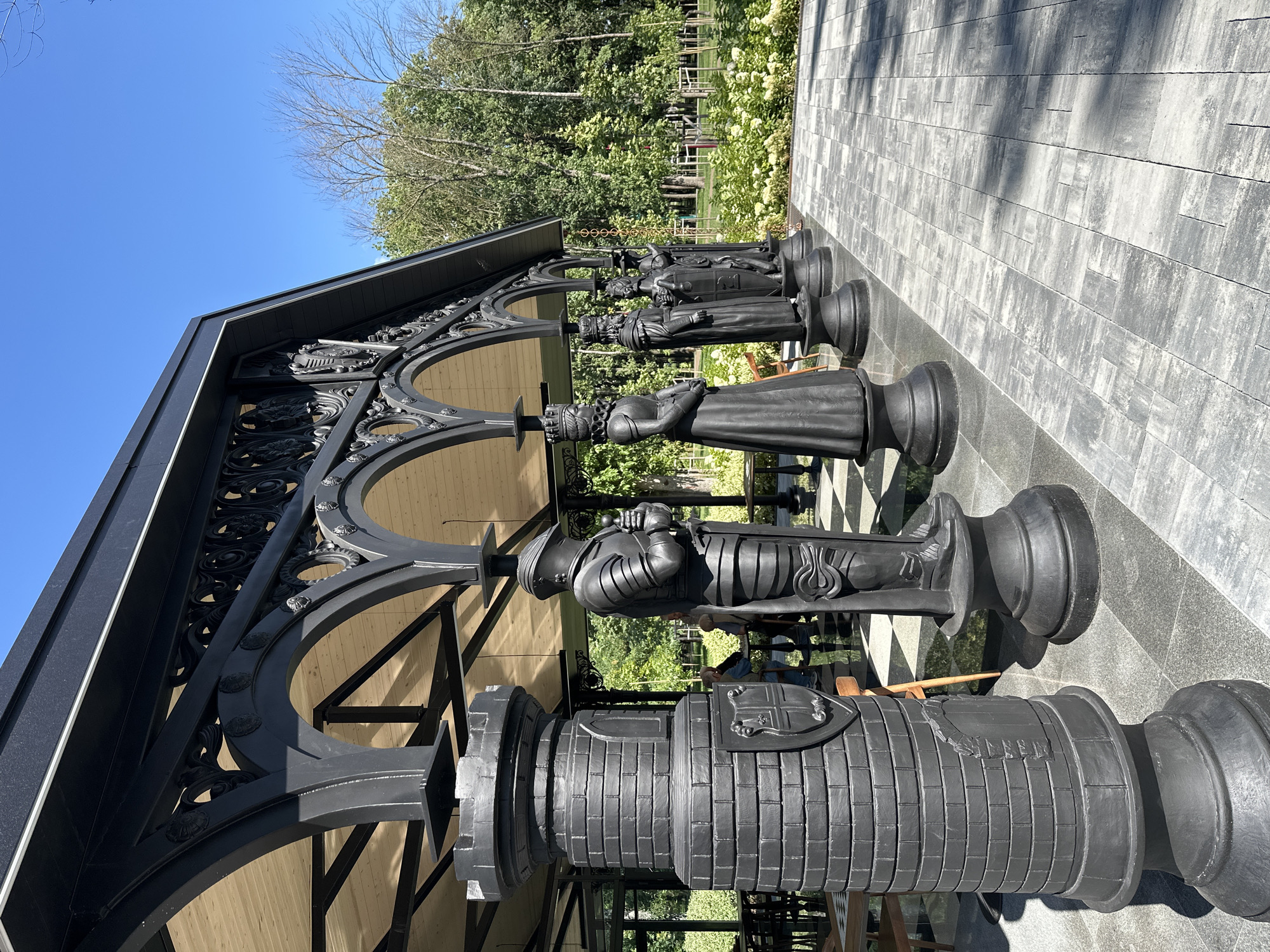 The image shows an outdoor covered seating area with a dark, ornate metal roof supported by columns. Several large, dark chess pieces are displayed along the covered walkway, including a rook, a knight in armor, and several figures resembling royalty. The chess pieces are positioned on a checkered floor. In the background, trees and greenery suggest a park or garden setting. Some tables and chairs are visible inside the covered area, and a few people are seated at them. The ground outside the covered area is paved with grey interlocking stones. The scene appears to be in a public outdoor space. The image was taken in Belgorod, Russia, at the "Belgorod Fortress" cultural complex.