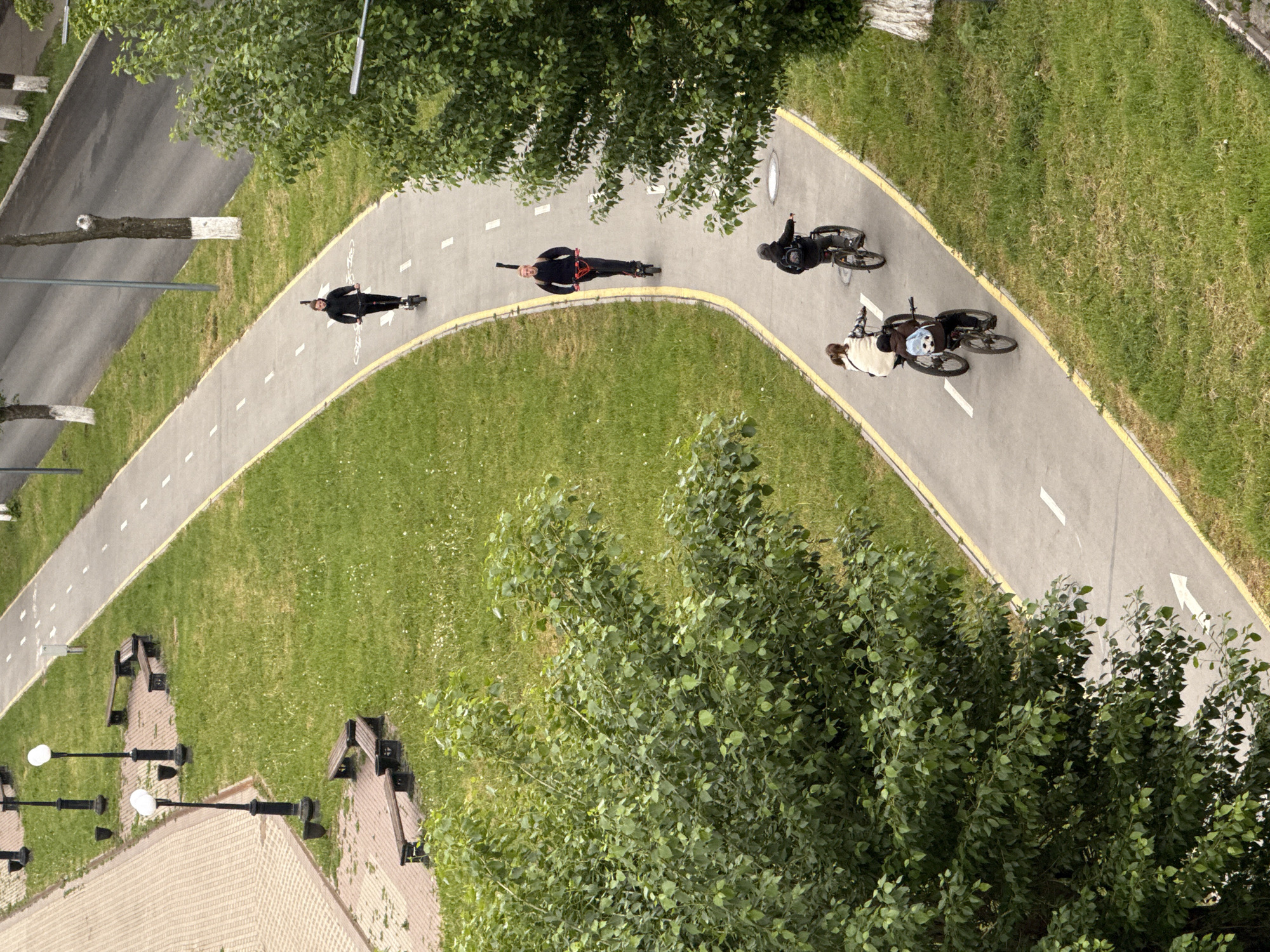 An aerial view shows a curved paved path in a green space. Four people are using the path. Two are on bikes, and two are on scooters. The path has a yellow border and white directional arrows. The green space is bordered by a road on one side. There are benches and lampposts visible near a building in the lower left corner. A row of trees is along the path and the road. This location is in the city of Votkinsk, Russia.