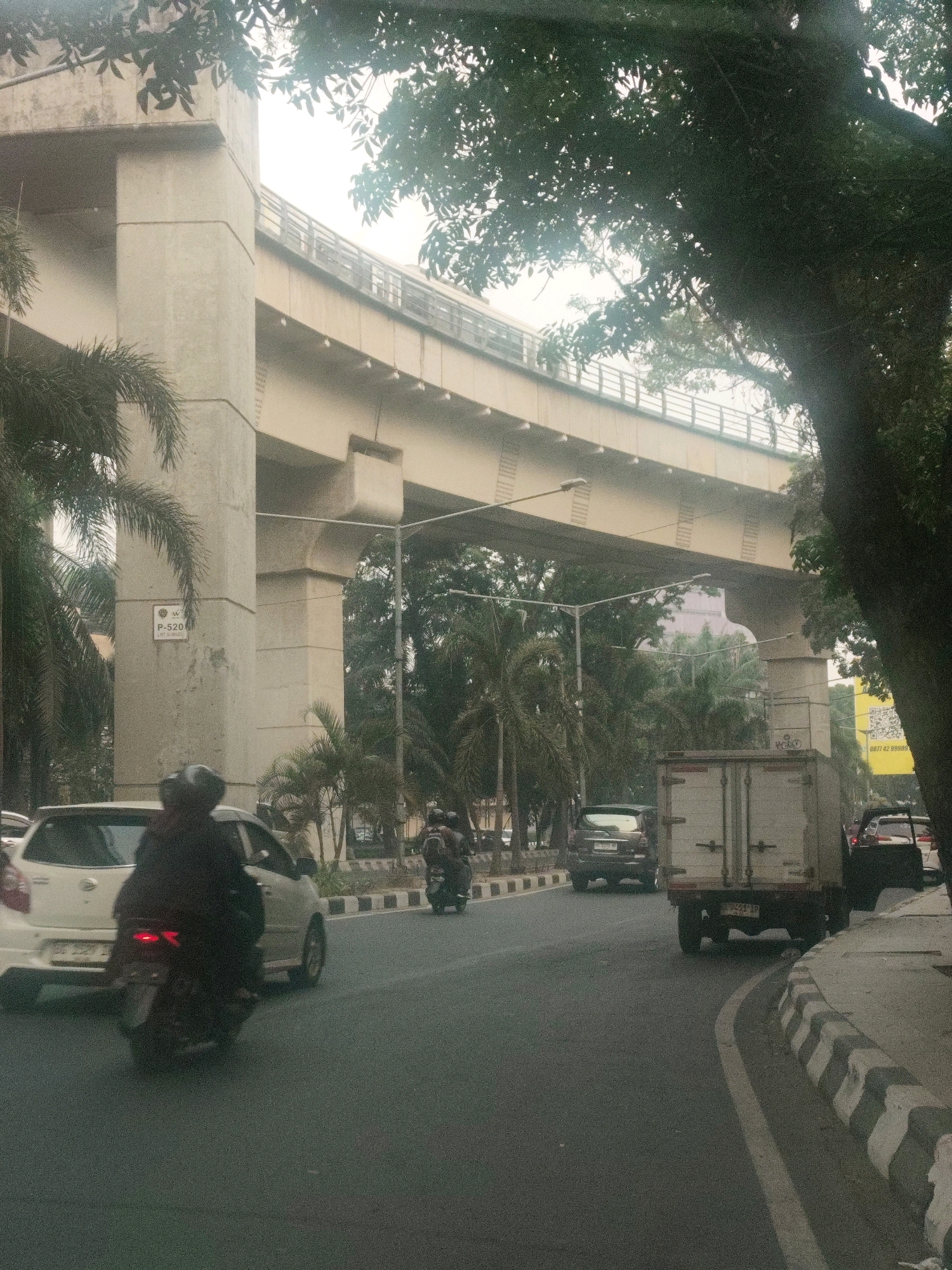 A street scene in Palembang, Indonesia, features an elevated highway structure with large concrete pillars supporting it. Cars and motorcycles are visible on the road. A white sedan is in the foreground on the left, followed by a motorcycle with a rider wearing a dark outfit. Further down the road, a light-colored truck with its rear doors closed is positioned in the right lane. Trees and foliage are present on the sides of the road, with a large tree in the foreground on the right partially obscuring the view. The sky appears overcast. A yellow banner with a QR code and text is visible on the right side of the frame.