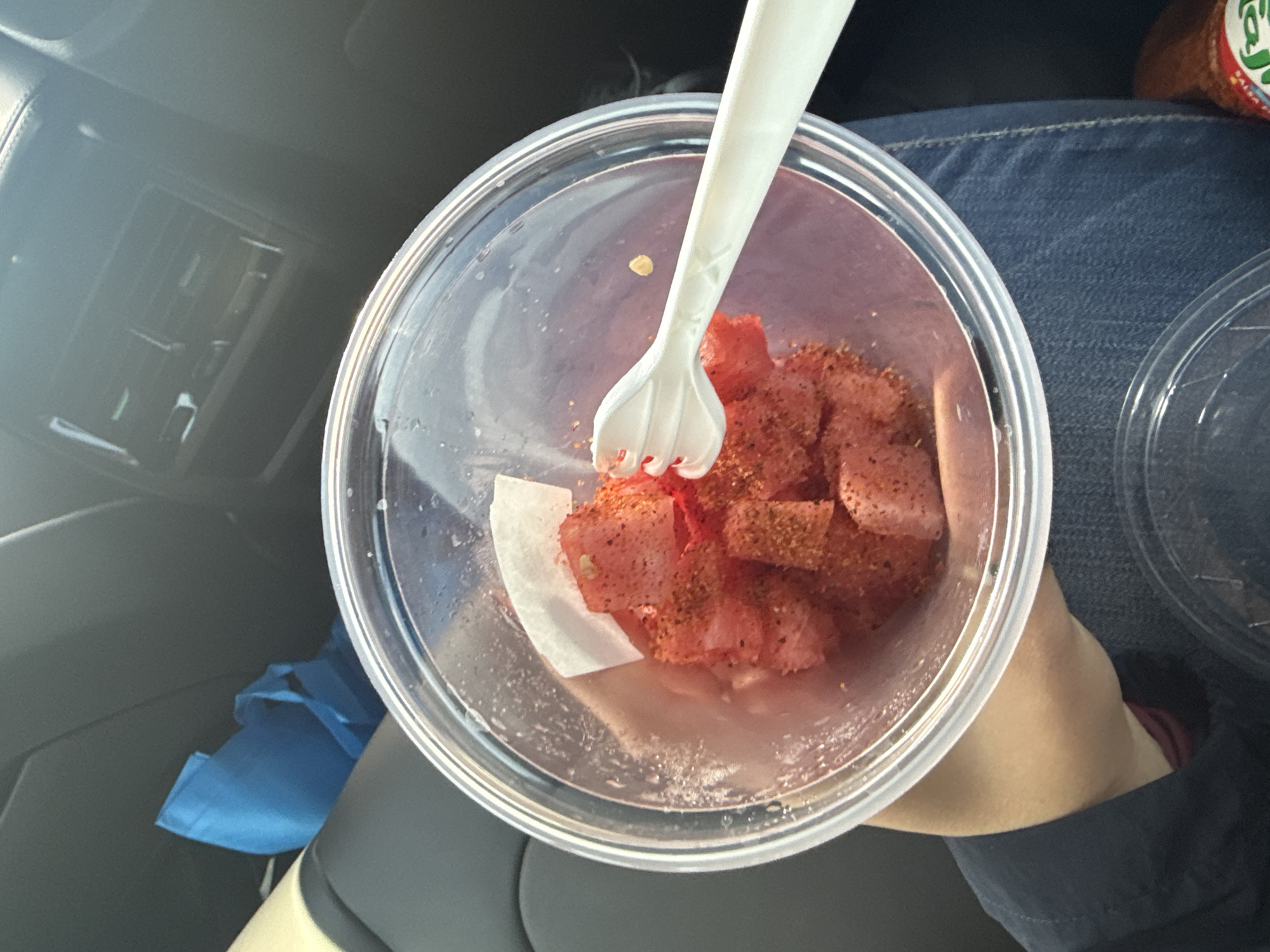 A top-down shot shows a plastic container filled with diced watermelon sprinkled with chili powder, held by a person wearing jeans and a dark jacket. A white plastic fork is stuck in the watermelon. The scene appears to be inside a vehicle, with a car's interior visible in the background. There is no evidence in the image that this is Ocotlán in the state of Jalisco, Mexico.