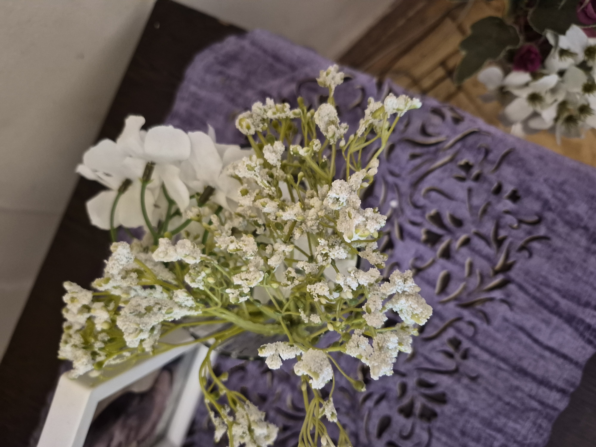 An overhead shot captures a static indoor arrangement of artificial flowers and decorative elements, likely on a table or shelf. The primary subject is a small bouquet of white artificial flowers. The most prominent are clusters of tiny, white, baby's breath-like blossoms, which have a distinctive textured, possibly frosted or glittery appearance. To the left of these, larger white artificial flowers with green stems are partially visible.
The floral arrangement is situated partly in front of a white picture frame, a corner of which is seen in the lower left. Beneath these items, a vibrant purple textile, likely a placemat or table runner, is spread across a dark brown wooden surface. This purple fabric features an intricate, dark, and ornate embossed or woven pattern. In the softly blurred background, another floral arrangement can be vaguely seen, containing white and dark pink or purple flowers with green leaves. There is no human activity depicted, nor is any text visible in the image. This scene was captured in Yerevan, Armenia.
