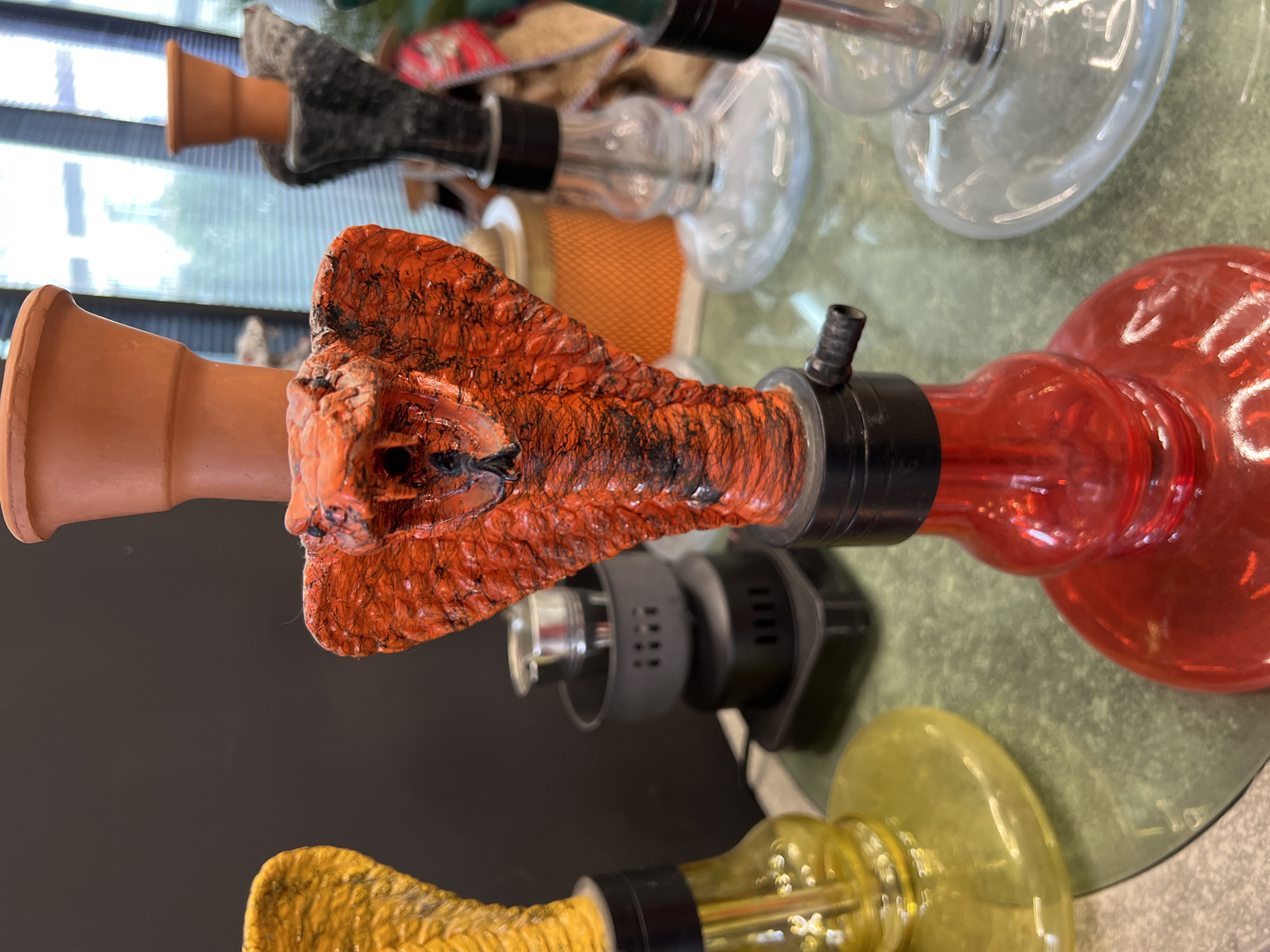 The image displays a close-up view of several hookahs, or shisha pipes, arranged on a surface, likely in Krasnodar, Russia. The central and most prominent object is a distinctive hookah with an orange, highly textured bowl, which appears to be sculpted to resemble a creature's head, possibly a snake or a stylized monster, complete with visible scales and dark, burnt-like accents. This unique bowl is attached to a terracotta-colored stem, which then connects to a translucent red glass base.
To the right and slightly above, another hookah is partially visible, featuring a greyish-black textured bowl, a similar terracotta-colored stem, and a clear glass base. Below the central orange hookah, part of a yellow glass hookah base can be seen. In the background, additional clear glass hookah bases are visible, suggesting a collection or inventory. All the hookahs are resting on a slightly reflective, greenish table surface, possibly glass or polished stone. Small black hose connectors are visible on some of the bases.
In the upper left background, horizontal blinds or window panes are discernible, indicating an indoor setting. The image captures the hookahs in a static display, with no people present or activity taking place. The lighting is consistent with an indoor environment. There is no visible text in the image.