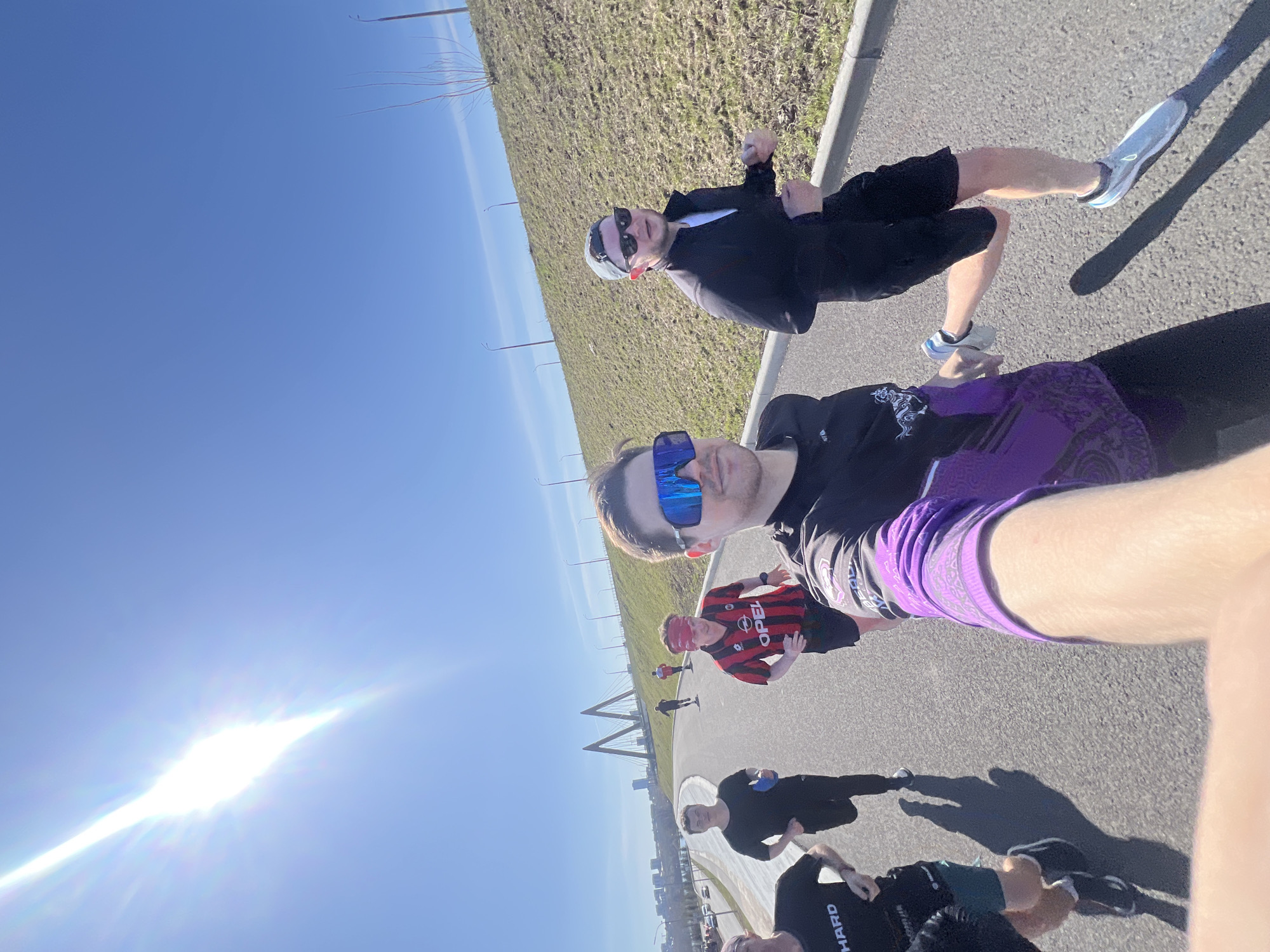 A group of people are walking on a paved path in Kazan, Russia. The sky is clear and blue, and the sun is shining brightly. The path is surrounded by grass and greenery. In the background, there is a bridge. There are several people walking on the path. They are wearing casual clothing and sunglasses. One person is wearing a red and black AC Milan jersey. One person is wearing purple shorts. The group appears to be taking a selfie. The location appears to be near the Millennium Bridge in Kazan, Russia.