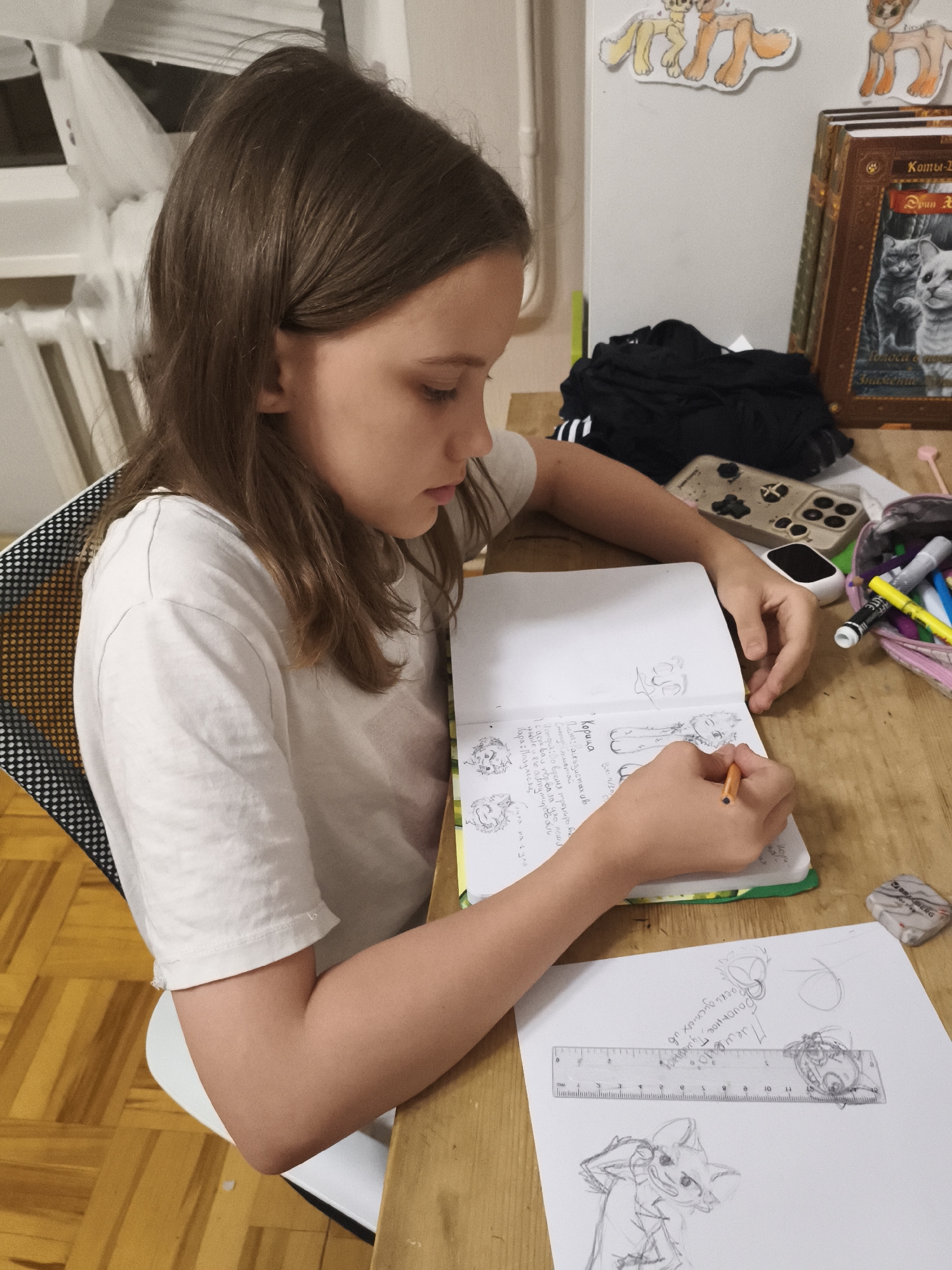 An indoor scene, likely a room within a residence, depicts a young girl focused on drawing and writing at a wooden desk. The girl, appearing to be pre-teen or early teen, has long, straight, dark brown hair and wears a plain white short-sleeved t-shirt. She is positioned in profile, looking down intently, holding an orange colored pencil in her right hand and actively working in an open notebook.
The notebook, open to a page with a green visible spine, contains sketches of cartoon characters/animals and handwritten text in Cyrillillic script. To the right of the notebook, on the light-colored wooden desk, is a separate white sheet of paper displaying additional sketches, including a detailed drawing of an anthropomorphic fox-like creature at the bottom and smaller sketches with Cyrillic text further up. A clear plastic ruler with black markings is placed horizontally across this paper. A grey and white eraser, partially labeled "BRAUBERG," rests on the far right of the desk. Adjacent to the girl's right hand is a pink/purple fabric pencil case filled with various colored pencils and markers, including a yellow marker with "LITE" visible text. A smartphone in a light-colored case with black embellishments, featuring a three-camera system, lies face down on the desk between the girl's left hand and the pencil case. A small white rectangular electronic device, possibly a earbud charging case, is next to the phone. Behind these items, a folded black t-shirt with white stripes, possibly Adidas branded, is casually placed on the desk.
The girl is seated on a chair with a black and white polka-dotted mesh backrest, visible on the left. The floor is covered in a light and dark brown parquet pattern. The background features a light-colored wall with two cut-out drawings of stylized cat-like figures, possibly from the "Warrior Cats" franchise, attached above the desk. To the right, two hardcover books are stacked vertically; the top book clearly displays "Коты-Воители" (Warrior Cats) and "Эрин Хантер" (Erin Hunter) in Cyrillic, with an illustration of two cats. A white-framed window with sheer white curtains or blinds is partially visible in the upper left background. The overall lighting suggests an indoor setting, possibly with artificial light.