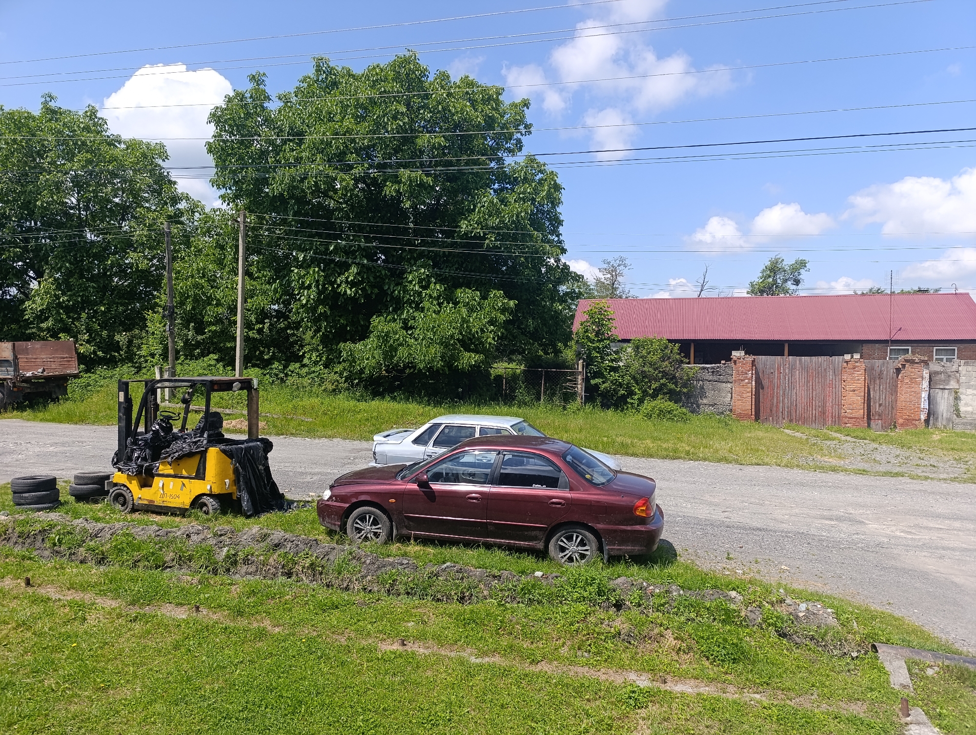 In Atabe, Georgia, under a bright, cloudy sky suggesting midday, a rural scene unfolds. In the foreground, a vibrant green lawn slopes upwards, bordered by a low, dirt embankment. To the left, a stack of old tires lies on the grass next to a yellow forklift, which is partially covered by a black tarp. The forklift bears Cyrillic text, "ДП-1504". Beyond the forklift, a dark red sedan is parked, its wheels turned slightly. Further back, a lighter colored sedan is parked behind it.
The middle ground features a gravel road that curves towards a large, red-roofed building with wooden double doors and brick accents. The building appears to be a barn or workshop. Behind the building, lush green trees dominate the landscape, their dense foliage reaching towards the sky. Power lines crisscross the upper portion of the image, a common sight in rural and semi-urban settings. The overall impression is one of a quiet, slightly overgrown rural area, possibly a farm or industrial yard on a sunny day. No people are visible in the image.
