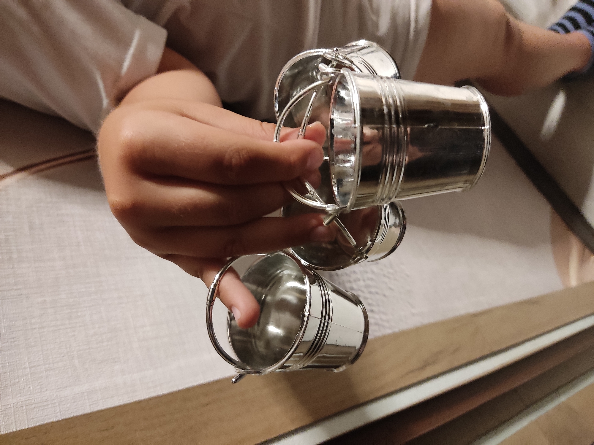 A person is holding three small, silver, metal buckets with handles. The buckets are shiny and reflective. The person's hand is visible holding two of the buckets, with a finger touching the inside of one. The background is out of focus, but appears to be indoors with a wall and possibly furniture. There is no information in the image to suggest this is in Noginsk, Russia.