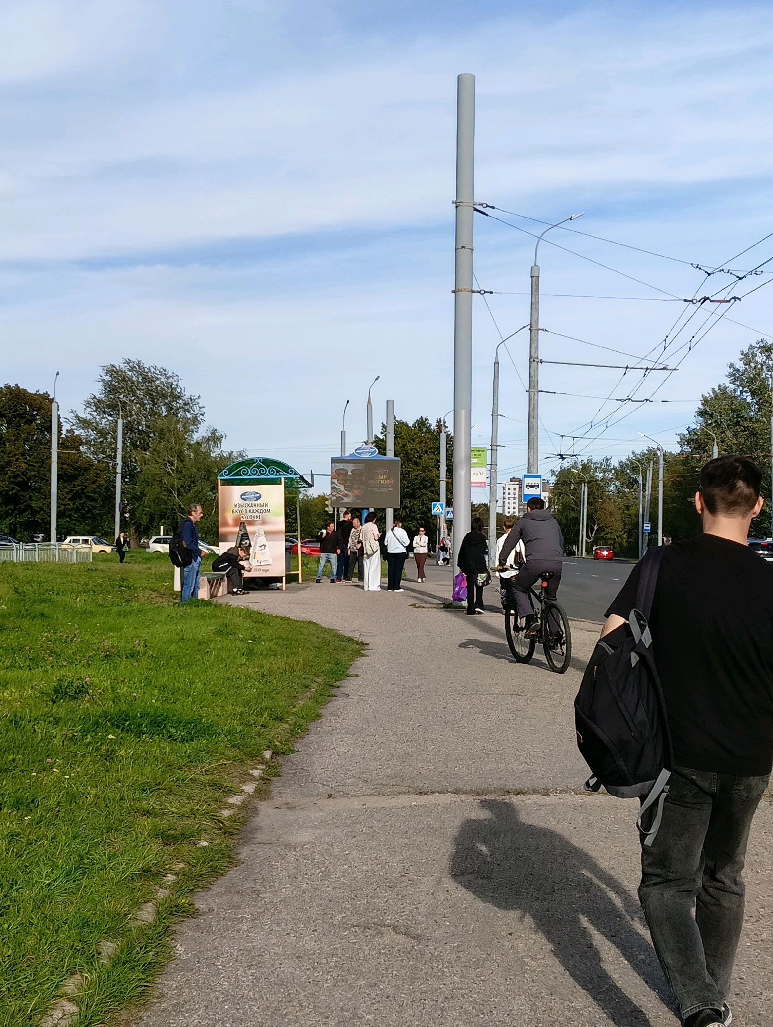 This is a ground-level, outdoor, full-shot photograph taken on a sunny day in the city of Hrodna, Belarus. The scene depicts a wide sidewalk with a grassy area to the left. Several people are visible, some walking, some waiting, and one person riding a bicycle. The background features trees, streetlights, and overhead electrical wires, suggesting a public transportation route, likely a trolleybus or tram line.
In the foreground, a person wearing a black t-shirt and dark jeans, carrying a black backpack, is walking away from the camera towards the right. Their shadow is cast on the pavement.
Further down the sidewalk, a person is riding a bicycle away from the camera. They are wearing a grey hoodie and dark pants.
Near the center of the image, there's a bus stop shelter with advertising posters. Several people are gathered around the bus stop, some standing and some sitting on a bench. One man in a blue shirt and jeans is facing away from the camera and appears to be talking to someone on the bench. Another group of people, some in light-colored clothing, are standing near the bus stop.
In the middle ground, there are more people walking along the sidewalk, and a pedestrian crossing sign is visible. Cars are present on the road to the right of the sidewalk.
The environment is urban, with a mix of green space and infrastructure. The sky is partly cloudy with blue patches. The overall atmosphere is one of daily urban activity.
