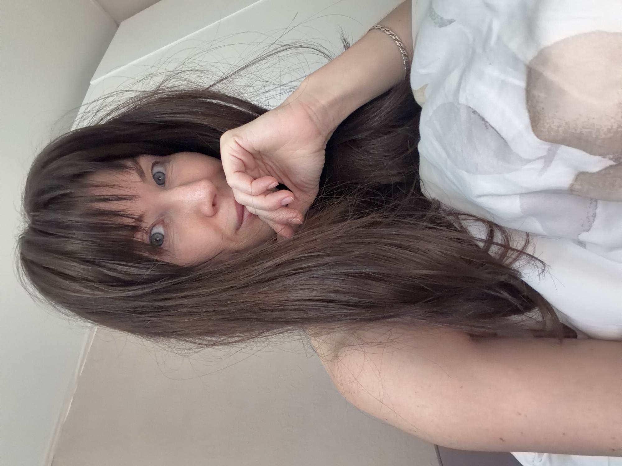 A woman with dark brown hair and bangs, fair skin, and blue eyes is lying in bed. Her head is tilted to the right, and her left hand is raised to her face, with her fingers curled slightly. She appears to be resting on a white pillow or bedding. Part of her right arm is visible. The background consists of a plain white wall and ceiling. This image does not depict any location in the city of Perm, Russia.