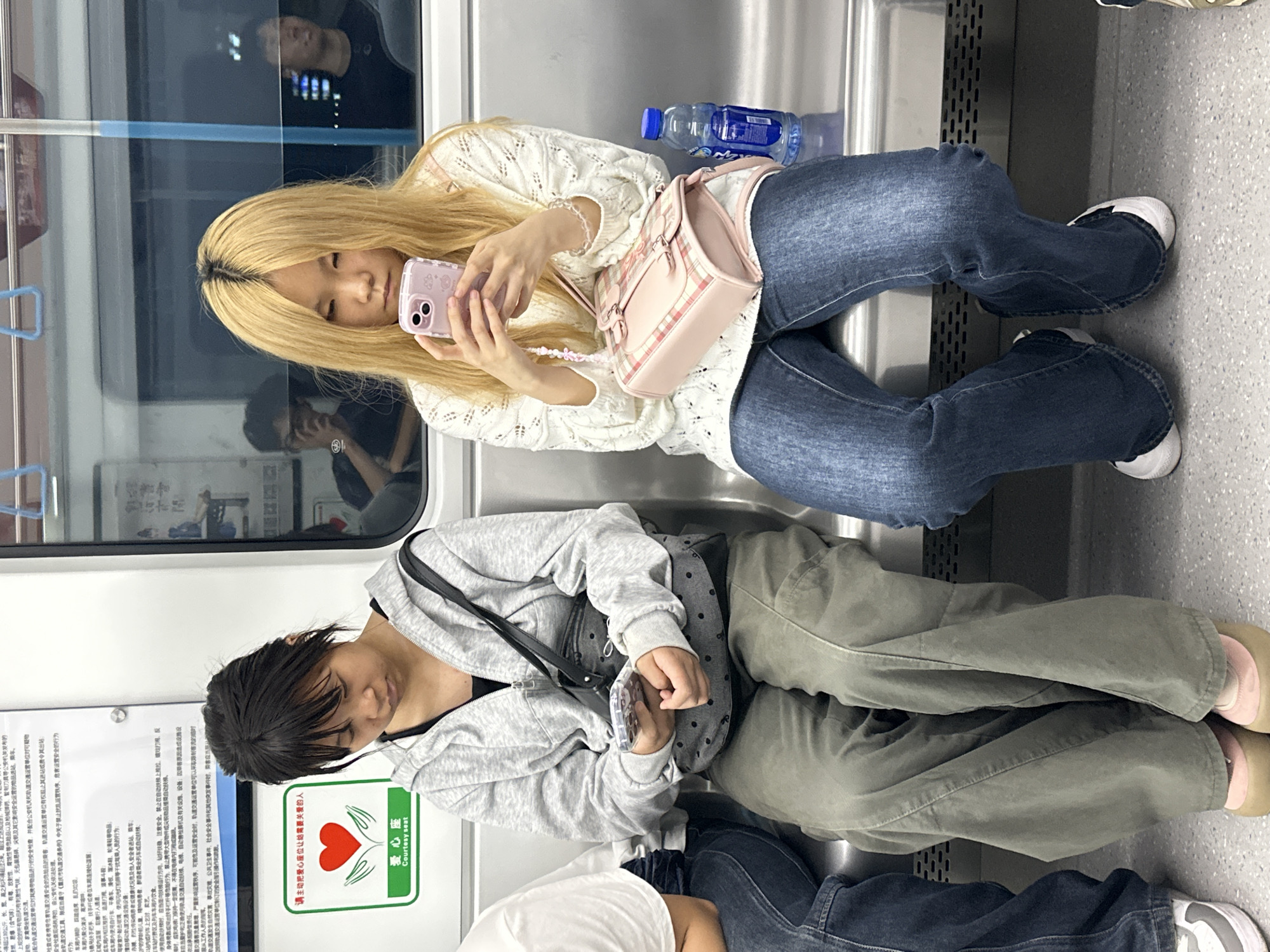 Two women are sitting on a train, seemingly in Tiaodeng, China. One woman has long blonde hair and wears a white top, blue jeans, and white shoes. She is holding a pink smartphone. She also has a pink purse and a bottle of water is nearby. The second woman has short dark hair and wears a grey hoodie, green pants, and tan shoes. She is also holding a smartphone and has a black purse. There is a courtesy seat sign nearby and a reflection in the train window. A third person is visible in the reflection of the train window.