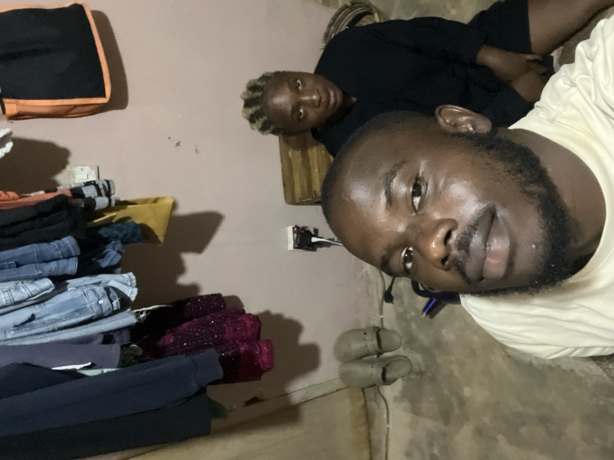 In Rafin Par, Nigeria, a close-up selfie captures a man and a woman in an indoor setting. The man, positioned in the foreground, is looking towards the camera with a beard and short hair. He is wearing a light-colored t-shirt. Behind him, a woman is seated and also looking towards the camera. She has braided hair and is wearing a black garment.
The background reveals a room with a textured, light-colored wall. Clothes are hanging on a rack, including denim jeans and other garments. A yellow bag and an orange and black bag are also visible on the wall. On the floor, there are two pairs of grey slip-on shoes. Electrical outlets and cords are present on the wall and floor. A wooden structure, possibly a stool or small table, is behind the woman. The floor appears to be concrete, with some texture and visible wires. The lighting suggests an interior environment with ambient light.