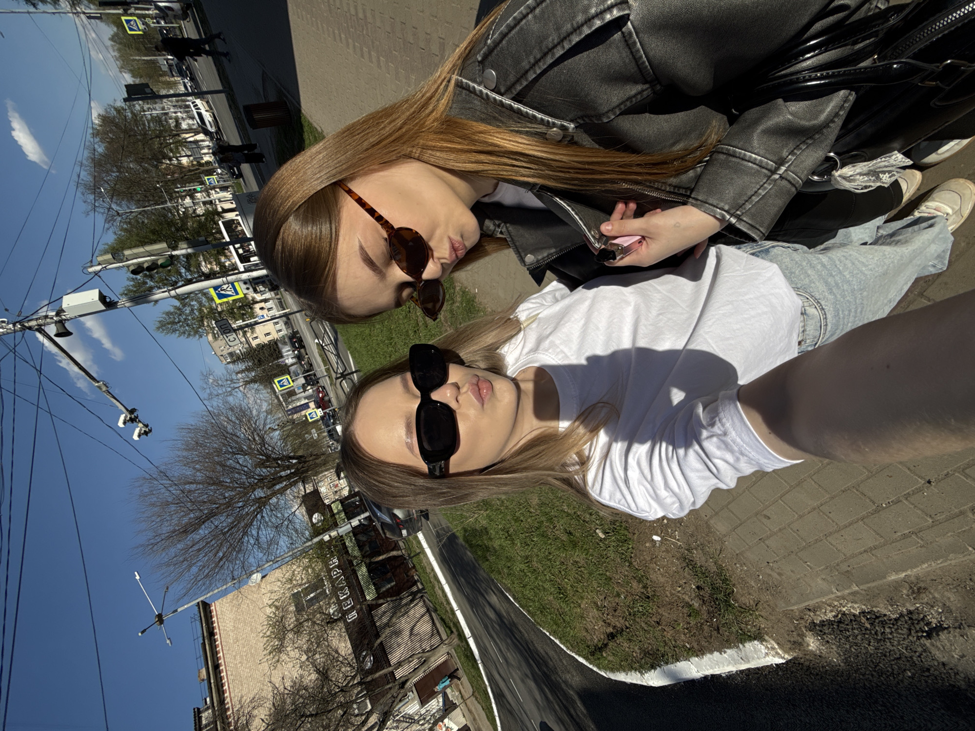 Two women are posing for a selfie outdoors. The woman on the top wears a black leather jacket, a white shirt, sunglasses, and light-colored pants. The woman on the bottom wears a white shirt, light blue jeans, and black sunglasses. Both are pouting their lips. They stand on a paved area with grass nearby. The location appears to be in the city of Chaikovsky, Russia, as evidenced by the sign visible in the background of the lower left-hand side which states "ЛЕКАРЬ," a medical term in Russian. The scene includes buildings, trees, power lines, street lights, and vehicles in the background. The weather is sunny.
