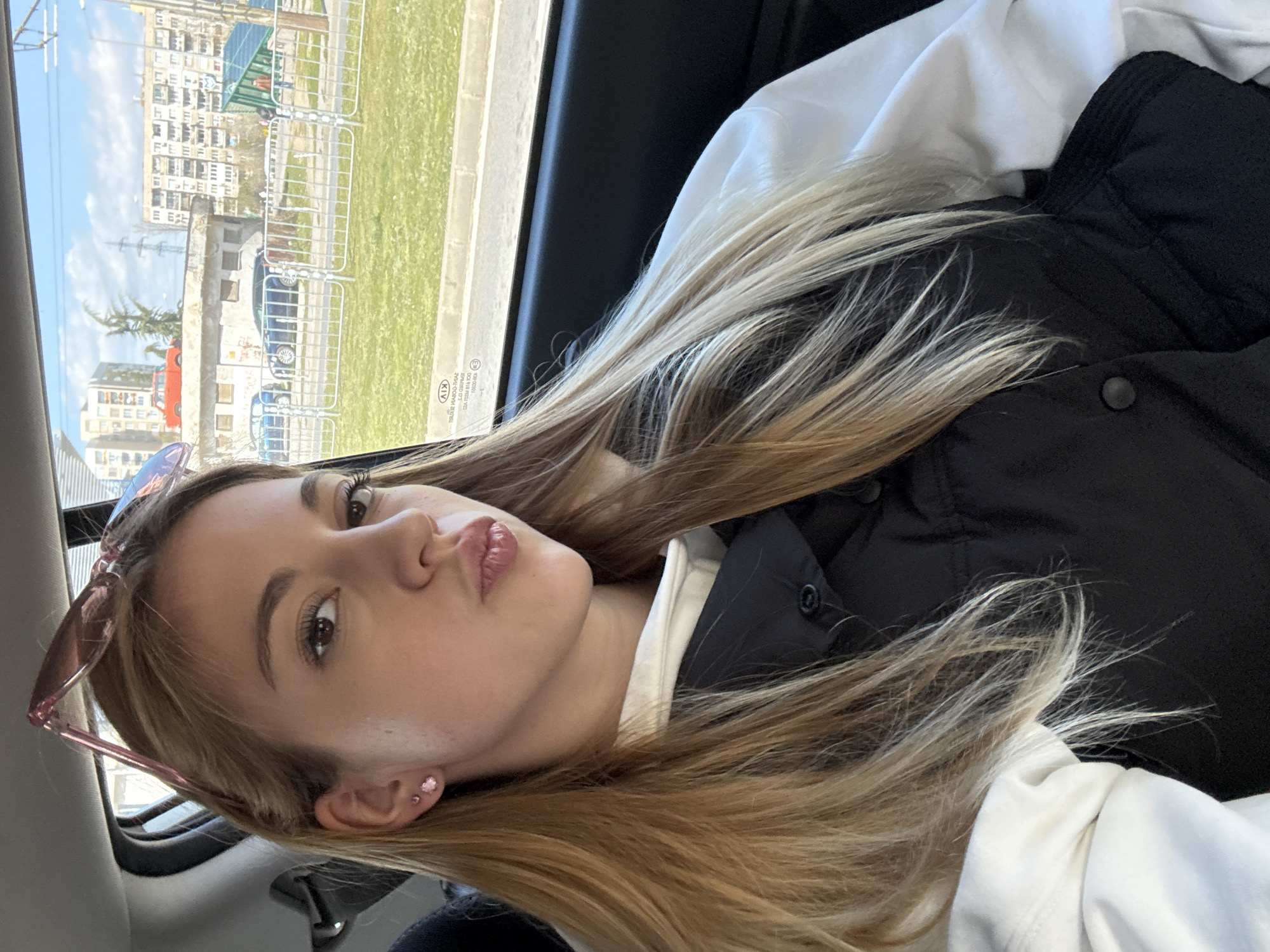 A selfie of a young woman is the central focus of the image, taken from a low angle inside a car. The woman has blonde hair and is wearing sunglasses on her head, a white sweatshirt and a black vest. She is pouting slightly, looking directly at the camera. The view through the car window reveals an urban landscape. Multi-story apartment buildings are visible across a grassy area, suggesting the setting is in Sofia, Bulgaria. There is a red car and other cars visible. Fences are present along the roadside.