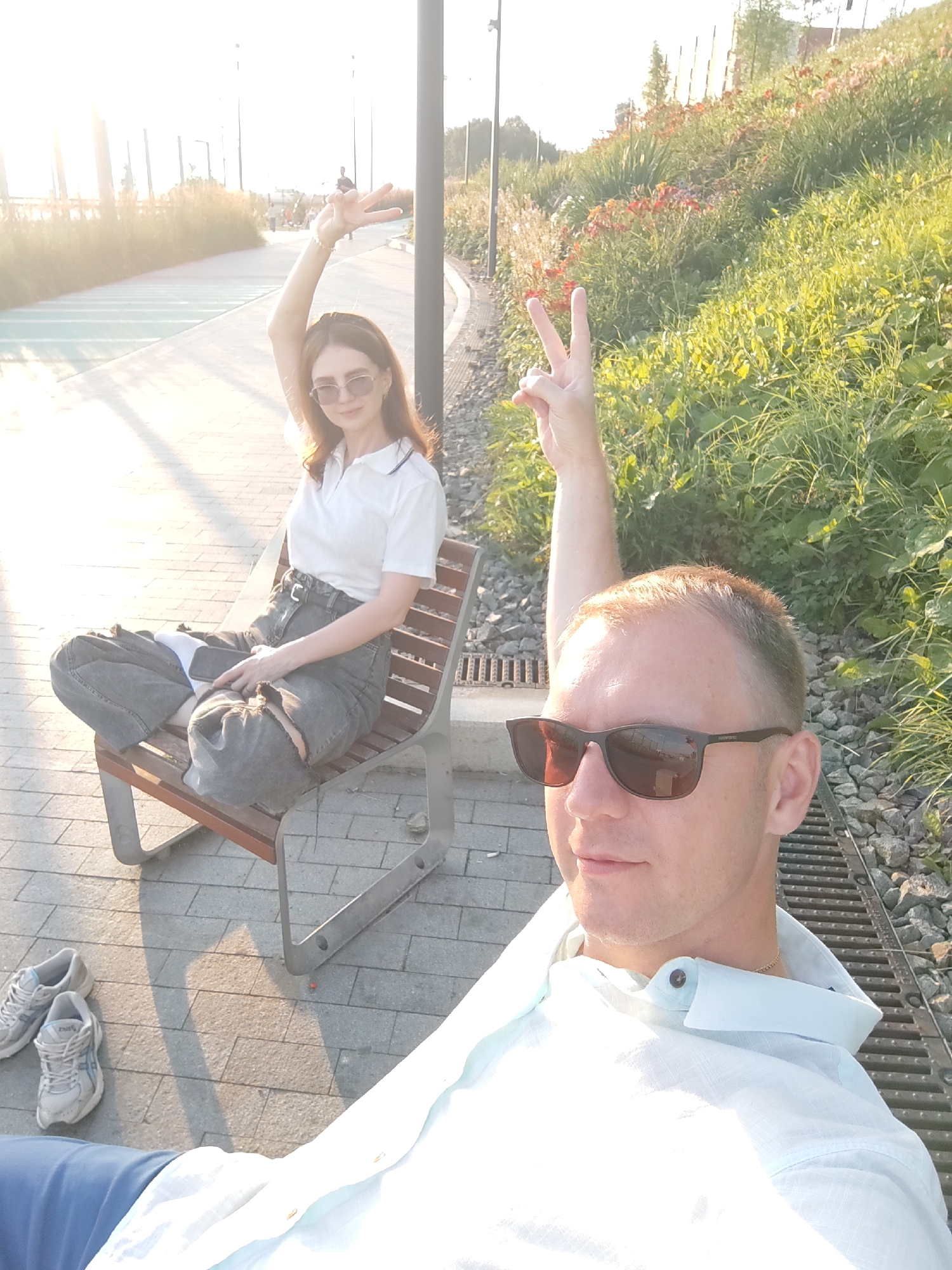 Two individuals, a male and a female, are pictured outdoors on a paved walkway next to a grassy embankment. The female is seated on a wooden bench, wearing a white collared shirt, dark ripped jeans, and sunglasses. She is raising her right arm and making a peace sign with her fingers. The male is in the foreground, taking a selfie, and is wearing a light blue collared shirt and sunglasses. He is also raising his right arm and making a peace sign. In the background, a pathway is visible with a light green section, suggesting a park or recreational area. The sun is shining, casting long shadows, indicating it is late afternoon. The presence of a pathway, landscaping with flowers, and streetlights suggests an urban park setting. The location is identified as the city of Perm, Russia.