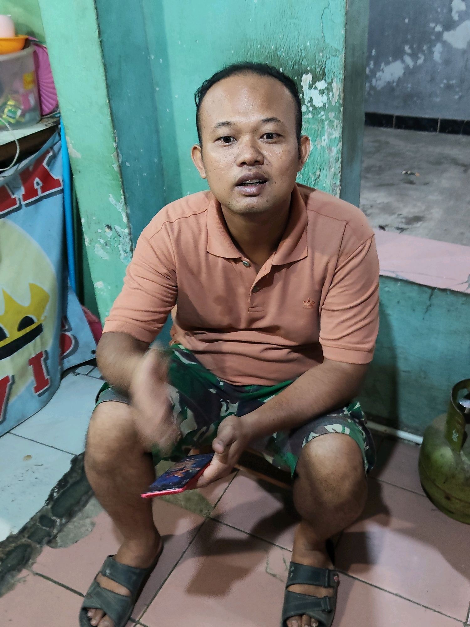 A middle-aged East Asian man with short dark hair and a light complexion is seated on a low stool in what appears to be an outdoor or semi-outdoor setting, possibly in Purwakarta, Indonesia. He is wearing an orange polo shirt and camouflage shorts. In his right hand, he holds a smartphone with a red case, displaying an image on the screen. His left hand is raised, palm open, as if gesturing. The background shows a weathered, teal-colored wall with peeling paint, and to the left, a blue and white banner with red lettering and a yellow graphic is partially visible. The floor is tiled with pinkish-brown and white tiles, and a portion of a green gas tank is visible on the right side of the frame. The lighting suggests it is daytime, and the overall scene conveys a casual, possibly domestic or informal commercial environment.