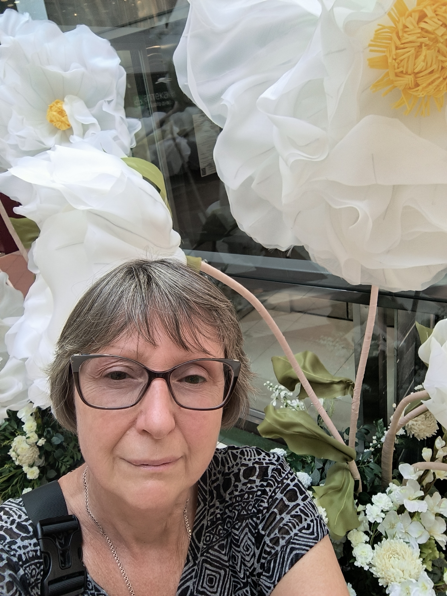 A medium close-up selfie is taken by a woman wearing glasses and a black and white patterned shirt. She is standing in front of large, decorative white flowers and a floral arrangement. The woman has short, graying hair and is looking directly at the camera with a slight smile. The background shows a glass storefront with some text visible, suggesting an indoor location, possibly a shopping center. The context of the city of Mykolaiv, Ukraine is not visually evident from the image itself, but the provided information states it is the location.