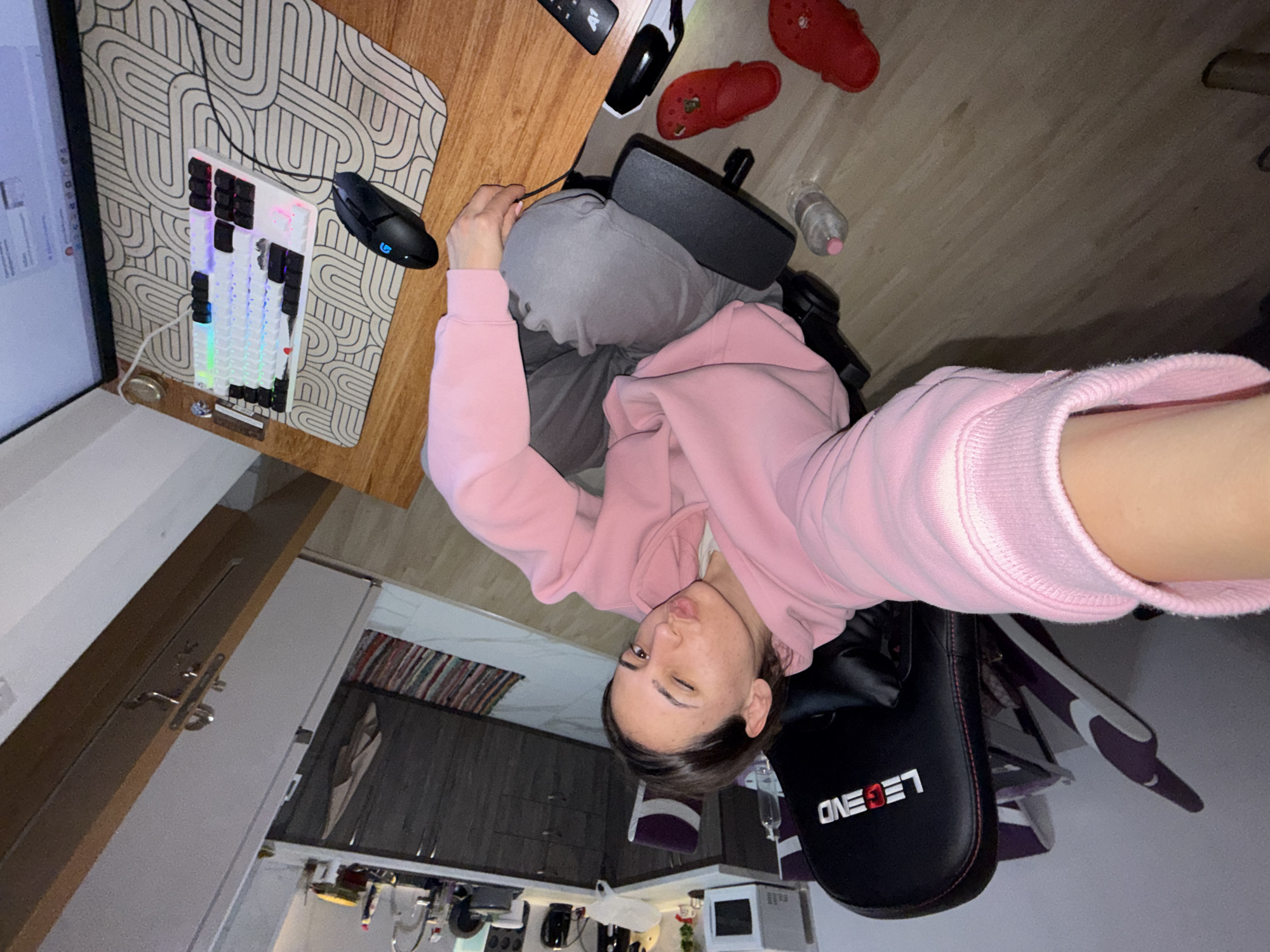 The image is a selfie taken by a person wearing a pink hoodie, sitting in a black and purple "Legend" gaming chair. The person is winking and appears to be in a kitchen area, as indicated by the cabinets, microwave, and countertop visible in the background. The person is in front of a desk with a computer monitor, keyboard, mouse, and a patterned mousepad. There are also red Crocs and a water bottle on the floor. There is no information that would enable to pinpoint a location in Plovdiv, Bulgaria.
