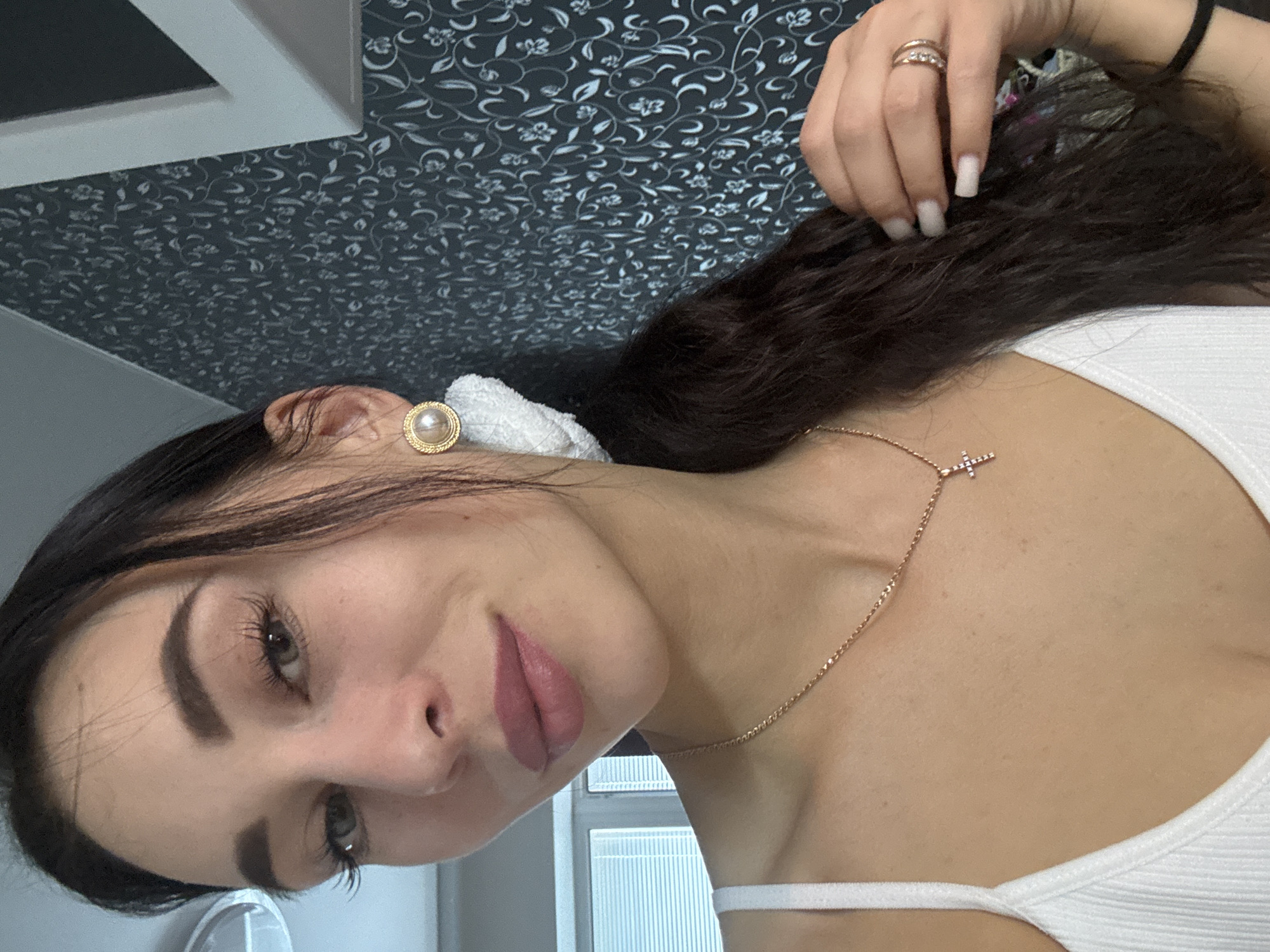 A woman with dark hair and fair skin is posing for a picture. She is wearing a white tank top, a gold necklace with a cross pendant, and gold pearl earrings. She has a ring on her right hand and appears to have long nails. The background consists of a patterned wall, a white framed object, and what seems to be a window. The location details, specifically Chaikovsky, Russia, cannot be determined from the visual information provided in the image.
