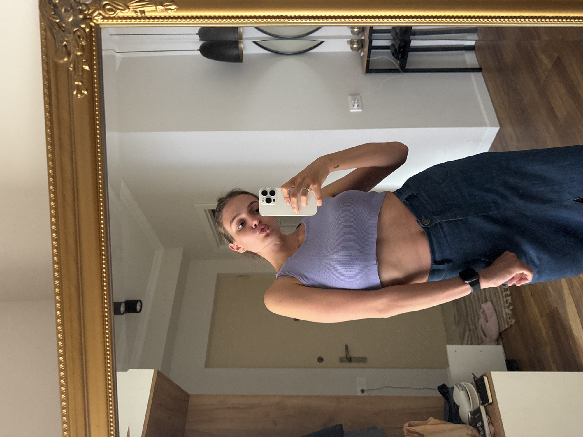A young woman is depicted taking a mirror selfie, with the image rotated 90 degrees clockwise. She appears to be standing, captured from just above her knees upwards. Her left arm is bent to hold a white smartphone, which covers the lower part of her face, leaving her eyes and forehead visible, along with a slight pout on her lips. She is wearing a light purple ribbed crop top that exposes her midriff and dark blue wide-leg jeans. A black smartwatch is on her right wrist, and a silver ring is visible on her left hand. A small tattoo reading "stay" is visible on her left forearm.
The selfie is taken in a large, ornate mirror featuring a thick, golden frame with intricate carvings and a beaded inner border. The reflection in the mirror shows an indoor setting with light-colored walls and a light brown wooden floor. Visible elements in the background include a black metal shelving unit, an electrical outlet, and a light-colored wooden door with a dark handle. Two distinct wall-mounted light fixtures, each featuring two black cylindrical lights, are reflected on opposite sides of the room. Near the bottom of the mirror, a white rectangular furniture piece, a light-colored rug, and some textile items on the floor are discernible. The room is brightly lit, indicative of an indoor daytime scene.