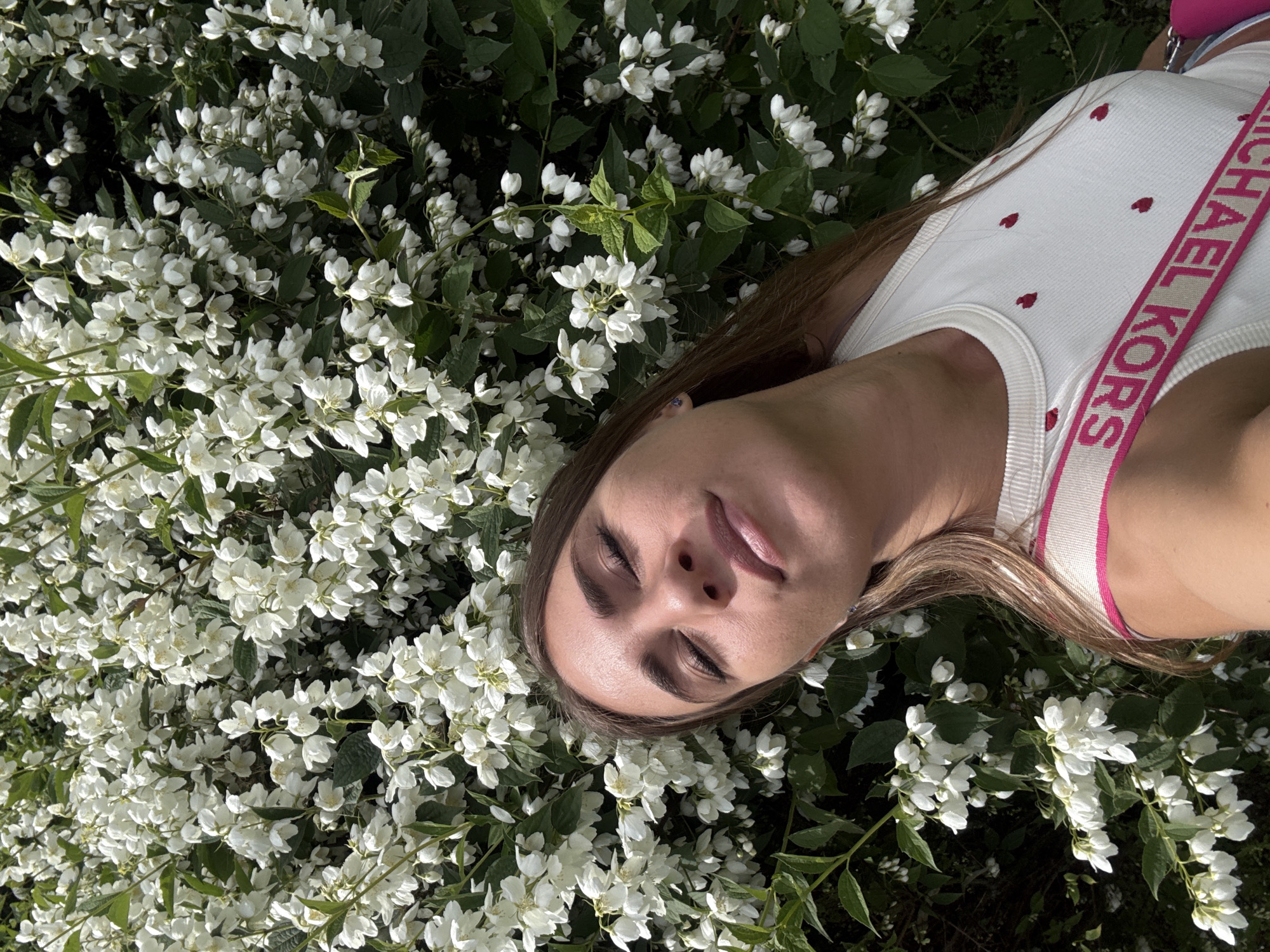 In this overhead, close-up shot, a young woman is lying amongst a dense bush of white flowers with green leaves. The woman is positioned diagonally across the frame, with her head near the center and her body extending towards the lower right. She has her eyes closed and her lips are slightly parted, suggesting a relaxed or peaceful state. Her long, light brown hair is spread out across the foliage. She is wearing a white tank top adorned with small red hearts, and a pink and white striped strap crosses over her shoulder, with the words "MICHAEL KORS" printed in pink on it. The lighting suggests it is daytime, possibly late afternoon, with dappled sunlight filtering through the leaves. The scene appears to be outdoors in a garden or park setting. The overall mood is serene and natural. The visible text on the strap identifies the brand of the accessory she is wearing. The presence of lush greenery and blooming white flowers indicates a warm season, likely spring or summer. Given the location context of Saint Petersburg, Russia, this scene likely takes place during the warmer months when vegetation is in full bloom.