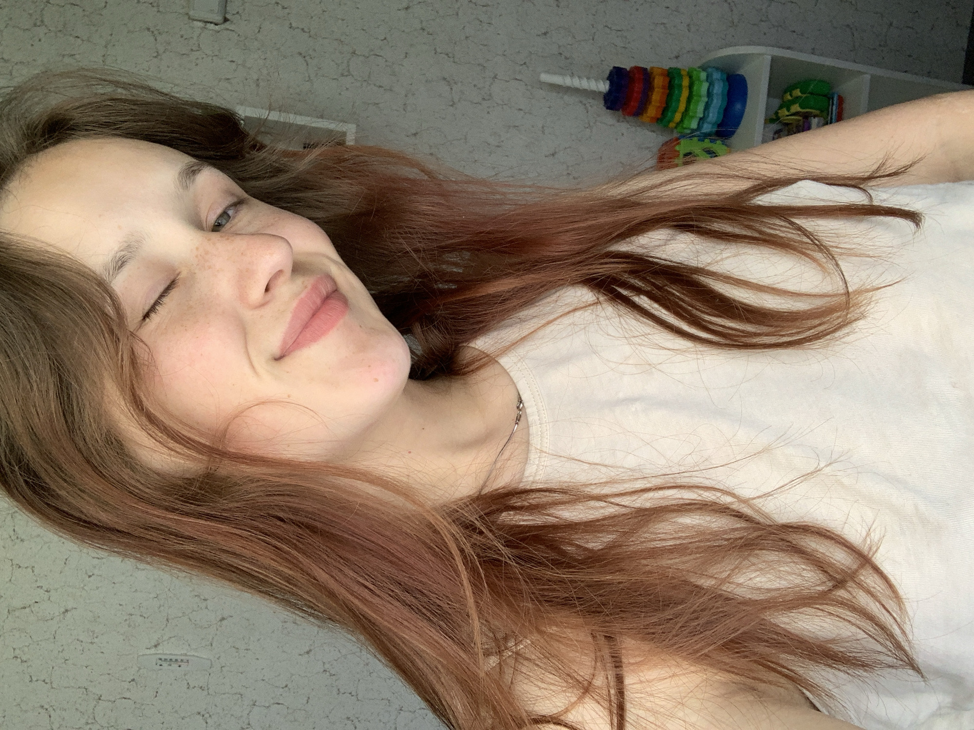 A young woman with reddish-brown hair is lying down, winking at the camera. Her skin is fair, with freckles on her nose and cheeks. She wears a light-colored shirt and a delicate silver necklace. In the background, there's a white shelf with colorful toys on top. The wall behind the shelf has a light grey pattern. There is no information available about the location of this image being in Фоки, Russia.