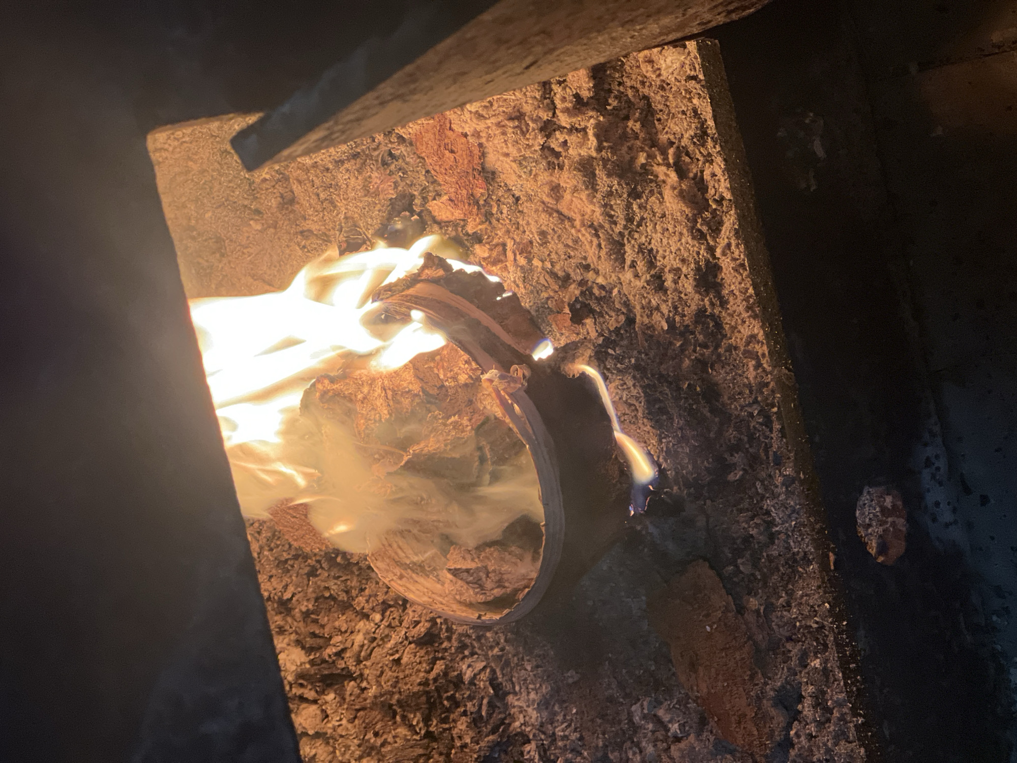 A contained fire burns within a wood-burning stove or fireplace. Flames engulf a section of what appears to be split wood or a similar combustible material. The surrounding interior of the stove is lined with a textured, heat-resistant material that is stained with soot and ash. The location, Chaikovsky, Russia, is not discernible from the image alone, but it is provided as contextual information. No people are visible.