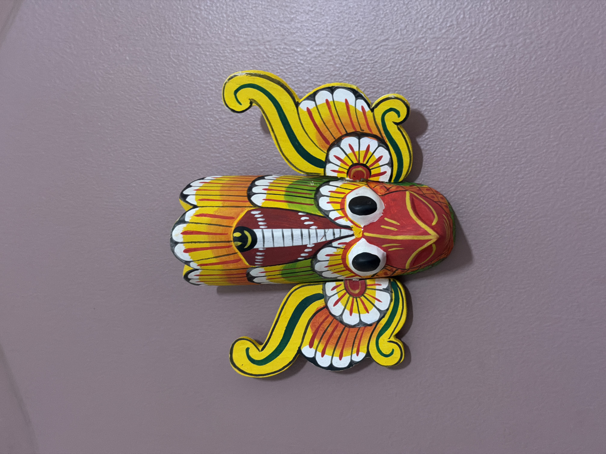 A vibrant, stylized wooden carving, resembling a mythical creature such as an owl or fish, is presented horizontally against a textured, light grayish-purple wall. The central body of the carving is elongated and segmented, painted with a vivid palette of red, yellow, green, white, and black. Two prominent, round black eyes with white surrounds are a key feature of the creature's head, which faces right.
Above and below the main body are two intricately designed, wing-like or fin-like extensions. These appendages are adorned with swirling yellow patterns, contrasting green stripes, and petal-like motifs in red and white, indicative of a folk art or traditional craft style. The surface of the carving shows some variations in paint application, revealing the texture of the wood beneath.
The wall behind the carving exhibits a subtle, stippled or stucco-like texture. Shadows cast by the object onto the wall suggest an overhead or upper-left light source, creating depth. No discernible activity is taking place; the image is a static portrayal of a decorative object. There is no visible text on the carving or the wall. The context places this scene in Moscow, Russia.