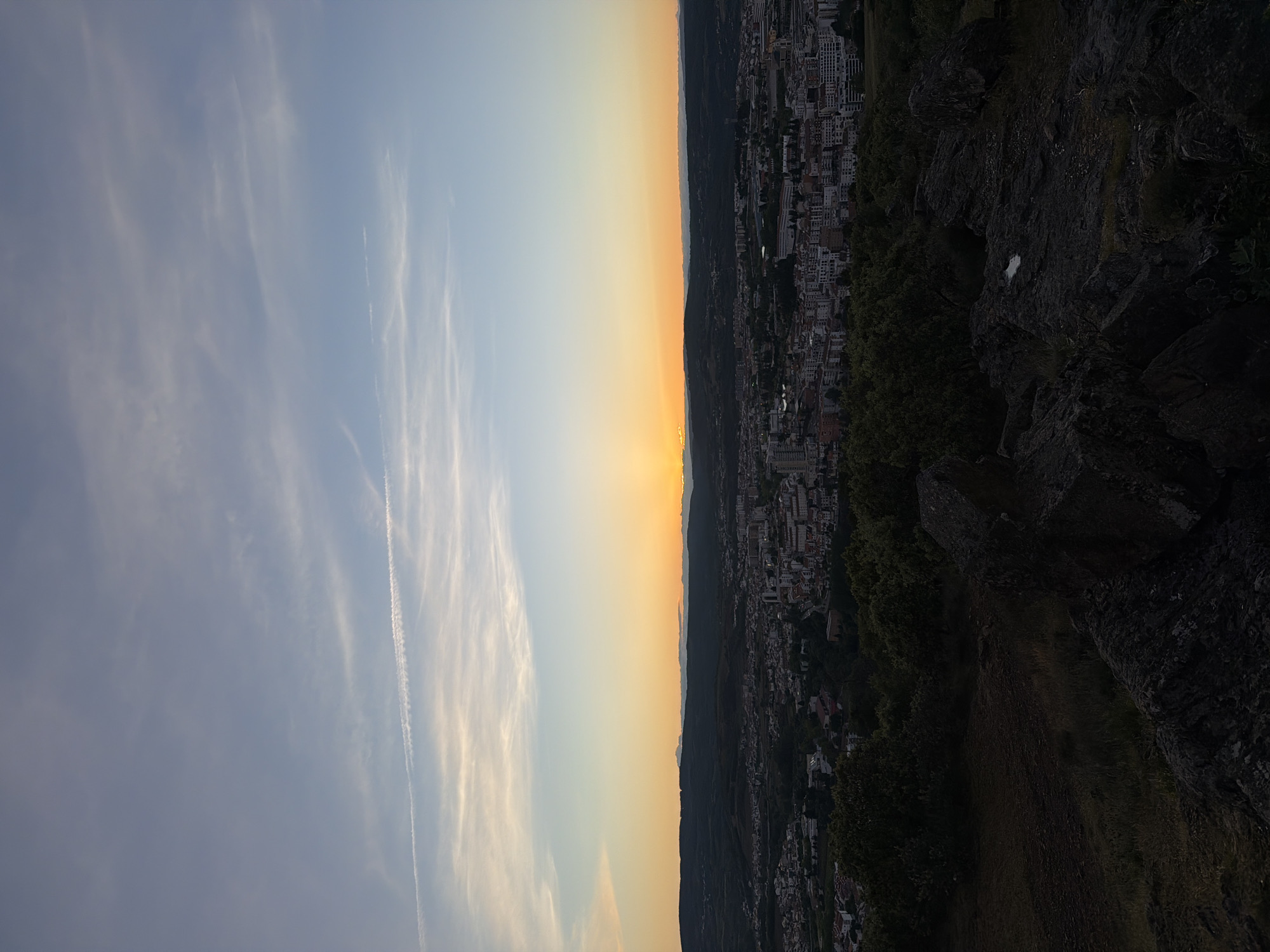 The image depicts a landscape overlooking the city of Bragança, Portugal, taken during sunset. The left side of the image shows a pale blue sky with wispy white clouds and contrails from aircraft. The sky transitions to a gradient of yellow and orange near the horizon, indicating the setting sun.
On the right side of the image, there is a view of Bragança. The city is nestled amidst hills and mountains. Many buildings are visible, including residential and commercial structures. The foreground of the right side features rocky terrain with vegetation. The image appears to be taken from a high vantage point overlooking the city.