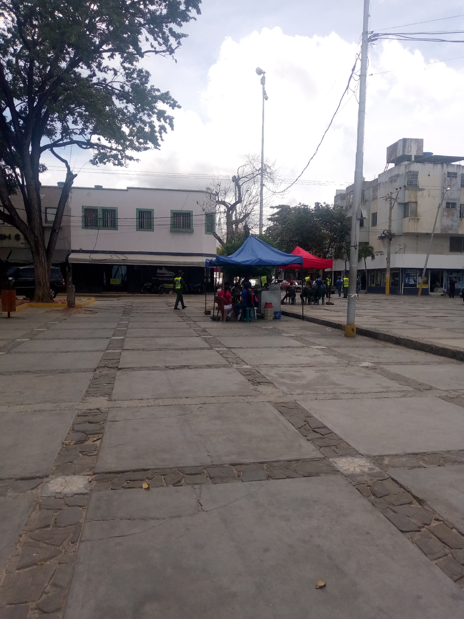 The image depicts a public square in Juan Griego, Venezuela. The square is paved with large concrete slabs divided by stone strips. In the background, there are two-story buildings with green-framed windows. A large tree is on the left, and power lines stretch across the sky. Several people are gathered under two canopies, one blue and one red. Some individuals wear yellow vests. There are benches and tables under the canopies, indicating a temporary gathering or event. The overall atmosphere is of a public space during daylight hours.