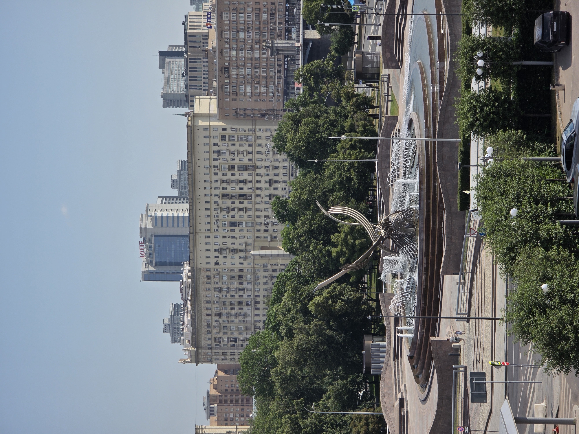 The image shows a wide shot of the central area of Moscow, Russia, on a clear day. In the foreground, there is a large, multi-tiered fountain with several jets of water spraying into the air. The fountain is surrounded by a curved stone plaza and walkway. Beyond the fountain, there is a dense area of green trees. In the background, several large buildings are visible. One prominent building is a light-colored, multi-story apartment building with many windows. To its left, a taller, modern building with a blue glass facade bears the "LOTTE" logo. To the right of the apartment building, a darker brick building with many windows is visible. Further in the distance, other urban structures and a bridge are discernible. In the mid-ground, two people, an adult and a child, are walking on the plaza near the fountain. The overall scene depicts a public space in Moscow, likely a park or square, with significant architectural elements and greenery.
