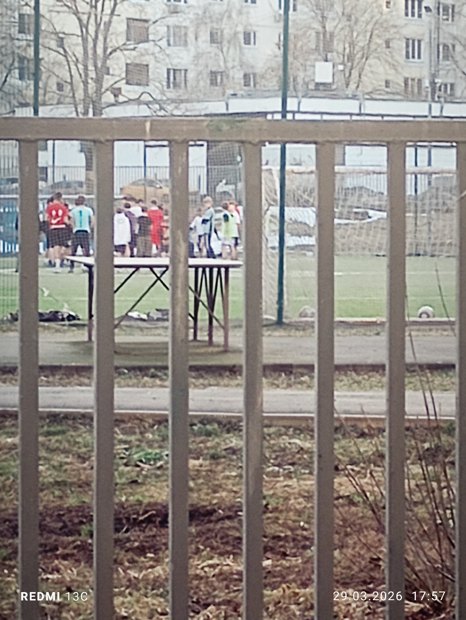 The image shows a fenced-off soccer field in Moscow, Russia. A group of people are gathered around a table on the field. Some individuals are wearing soccer jerseys. A ball is visible on the field. In the background, a multi-story building is visible. The fence is in the foreground, slightly obstructing the view. The photograph was taken on March 29, 2026, at 17:57.