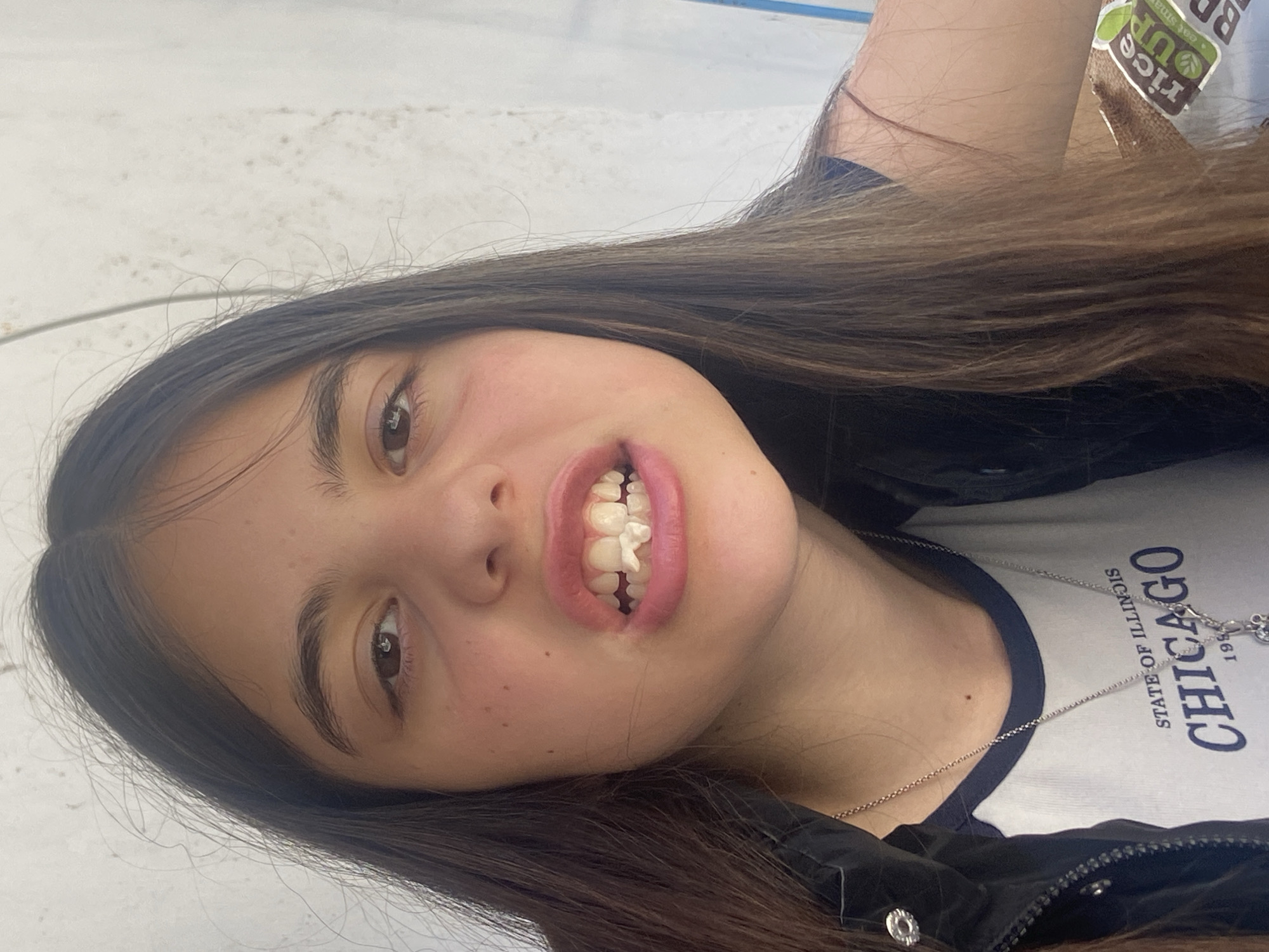 A young woman is posing for a photo. She has long brown hair, brown eyes, and is wearing a white t-shirt with "Chicago State of Illinois 199_" printed on it, along with a black jacket and silver necklace. She is smiling and has white material between her front teeth. The background appears to be a light-colored, possibly concrete or painted surface. Based on the available information, no specific location in Dragovishtitsa, Bulgaria, can be determined.