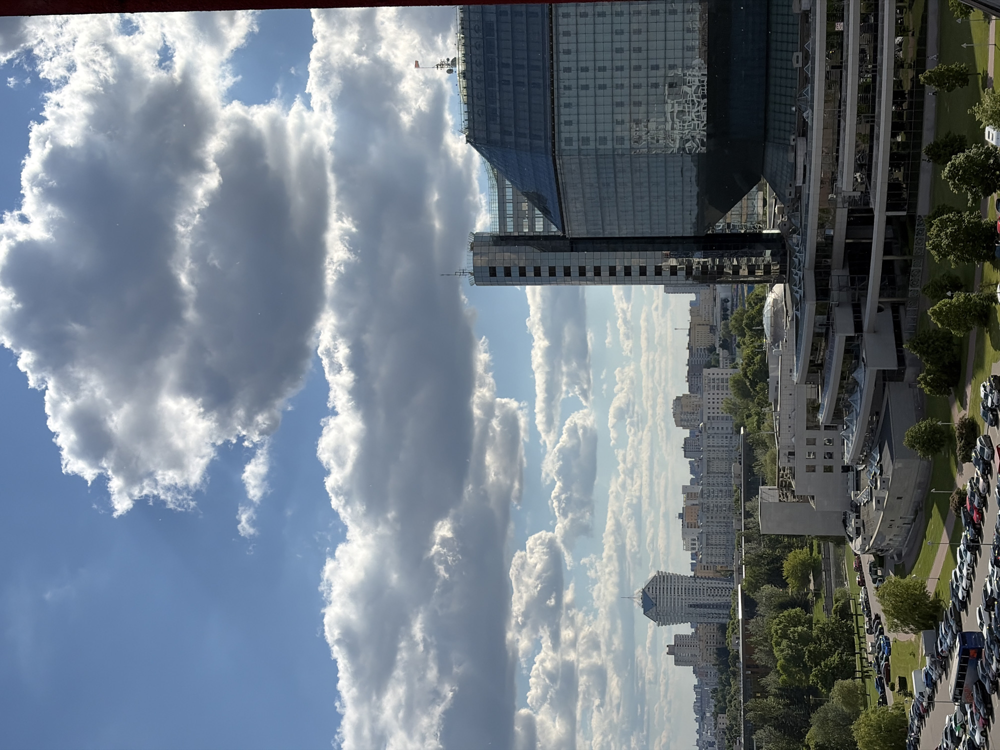 An eye-level, long-shot photo shows a panoramic view of Бараўлянскі сельскі Савет, Belarus on a sunny day. A building constructed primarily of glass features architectural designs with straight lines, angled planes, and a curvilinear shape that partially conceals the sky from a rooftop perspective. Behind the glass-fronted structure, several buildings dot the horizon, some of which have peaked rooftops. Immediately adjacent to the large glass building is a parking lot filled with cars of different colors, including many shades of blue, gray, and white. There are trees scattered throughout the landscape, with the most visible ones in the foreground bordering the parking lot. In the sky, large clusters of puffy white clouds cover the blue sky, with the most significant cloud mass to the left of the photo. A red beam of unknown origin appears at the very top edge of the frame.
