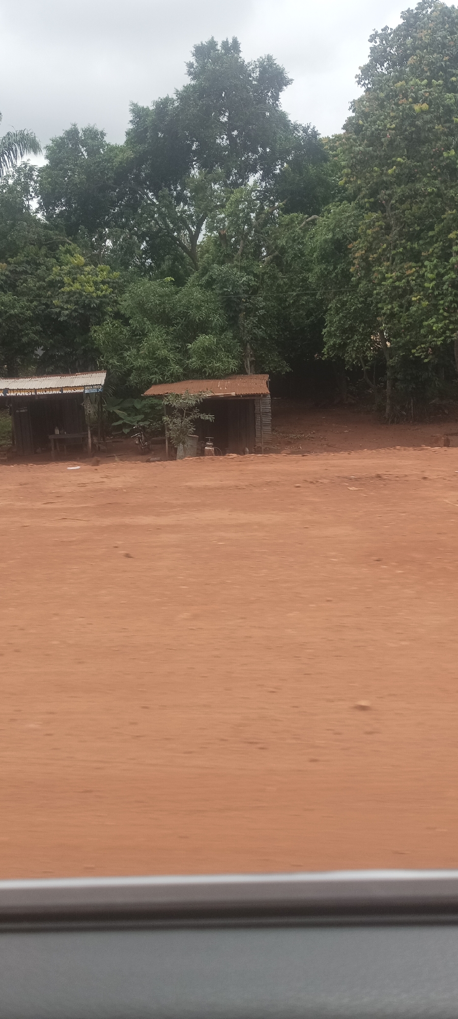 The image shows a dusty, reddish-brown dirt road in Imilike, Nigeria. Two small, open-air structures with corrugated metal roofs and wooden supports stand near the road. The structure on the left has a sign reading "JA JAPANESE MECHANIC WORKSHOP". The other structure contains tools and equipment, likely for automotive repair. Lush green trees and vegetation form a dense backdrop behind the structures. The sky is overcast. A car window is visible at the bottom of the frame, suggesting the photo was taken from a vehicle. There are no people visible in the frame.