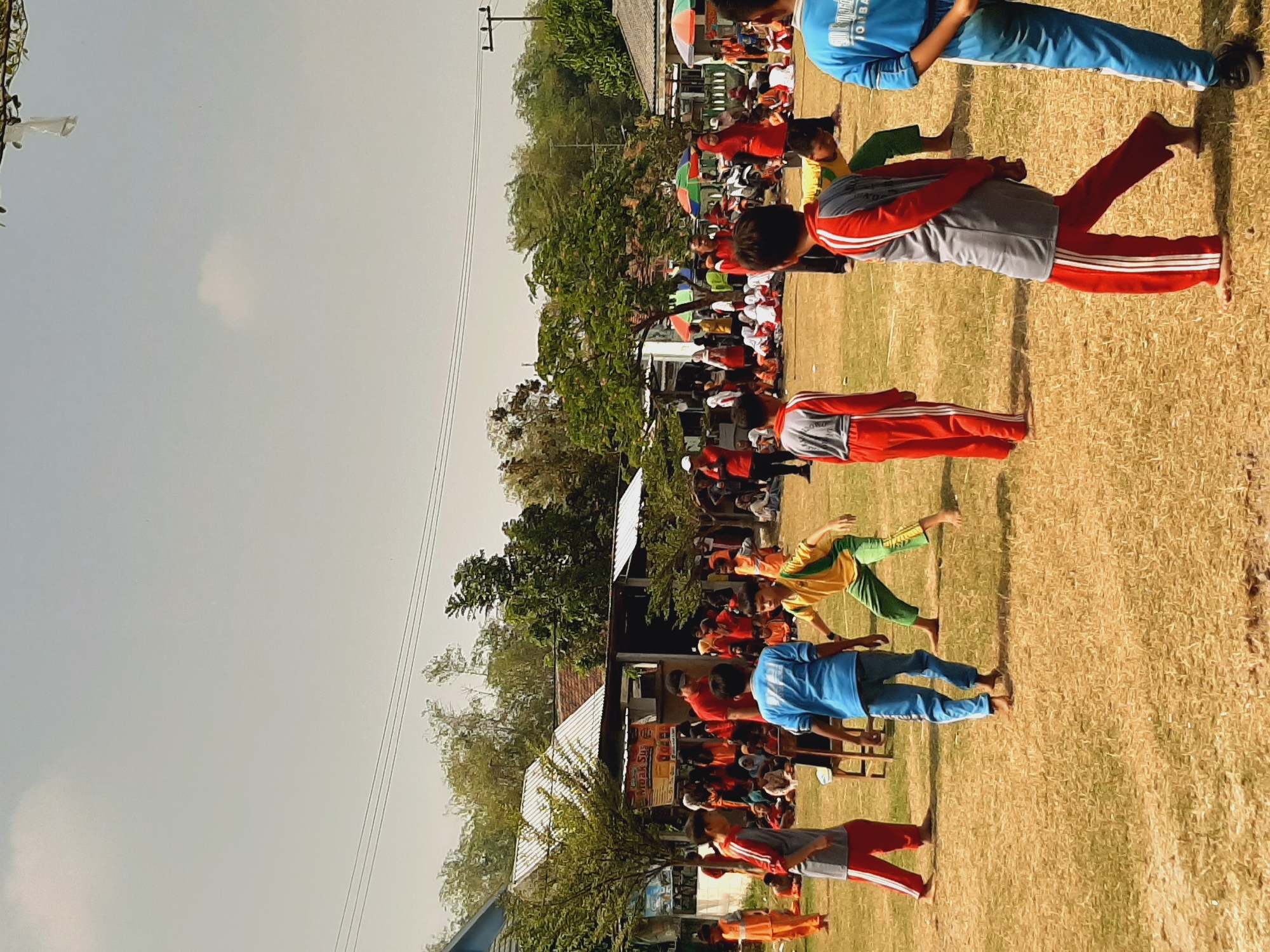 The image depicts an outdoor event, likely a school or community gathering, taking place on a dry, grassy field in Sumberjo, Indonesia. Several children, most of whom appear to be of elementary school age, are participating in activities. Some children are wearing athletic attire, including tracksuits in various colors such as red, blue, green, and yellow. One group of children appears to be engaged in a game or competition, with one child in a yellow and green outfit in motion, and another in a blue tracksuit standing nearby. Other children are seated on the ground, watching the events or resting.

In the background, there are several buildings with white, sloped roofs, suggesting a school or community center. Trees with green foliage are scattered around the area, and some colorful umbrellas are visible, providing shade. A sign with text, partially obscured, hangs on one of the structures. A few adults are also present, some of whom appear to be supervising or participating in the event. The sky is overcast with a pale, hazy appearance. The overall scene conveys a sense of active engagement and community participation.