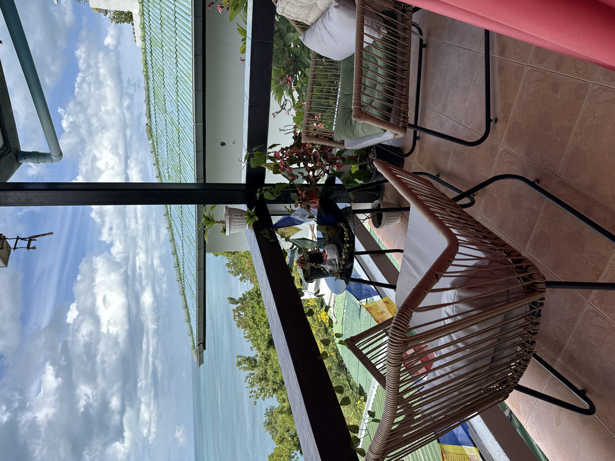 On a covered balcony in Ko Samui District, Thailand, two wicker chairs with metal frames face out towards a scenic ocean view under a partly cloudy sky. The chair in the foreground is empty and has a white cushion. The chair on the right is occupied by a beige cushion. A small round black table between the chairs holds a small decorative cat figure, prayer flags, and a potted plant. More potted plants are visible on the railing and in the background. The balcony floor is tiled, and a portion of a pink curtain is visible on the right edge of the frame. Green rooftops and lush vegetation are visible below the balcony, leading to the blue ocean.