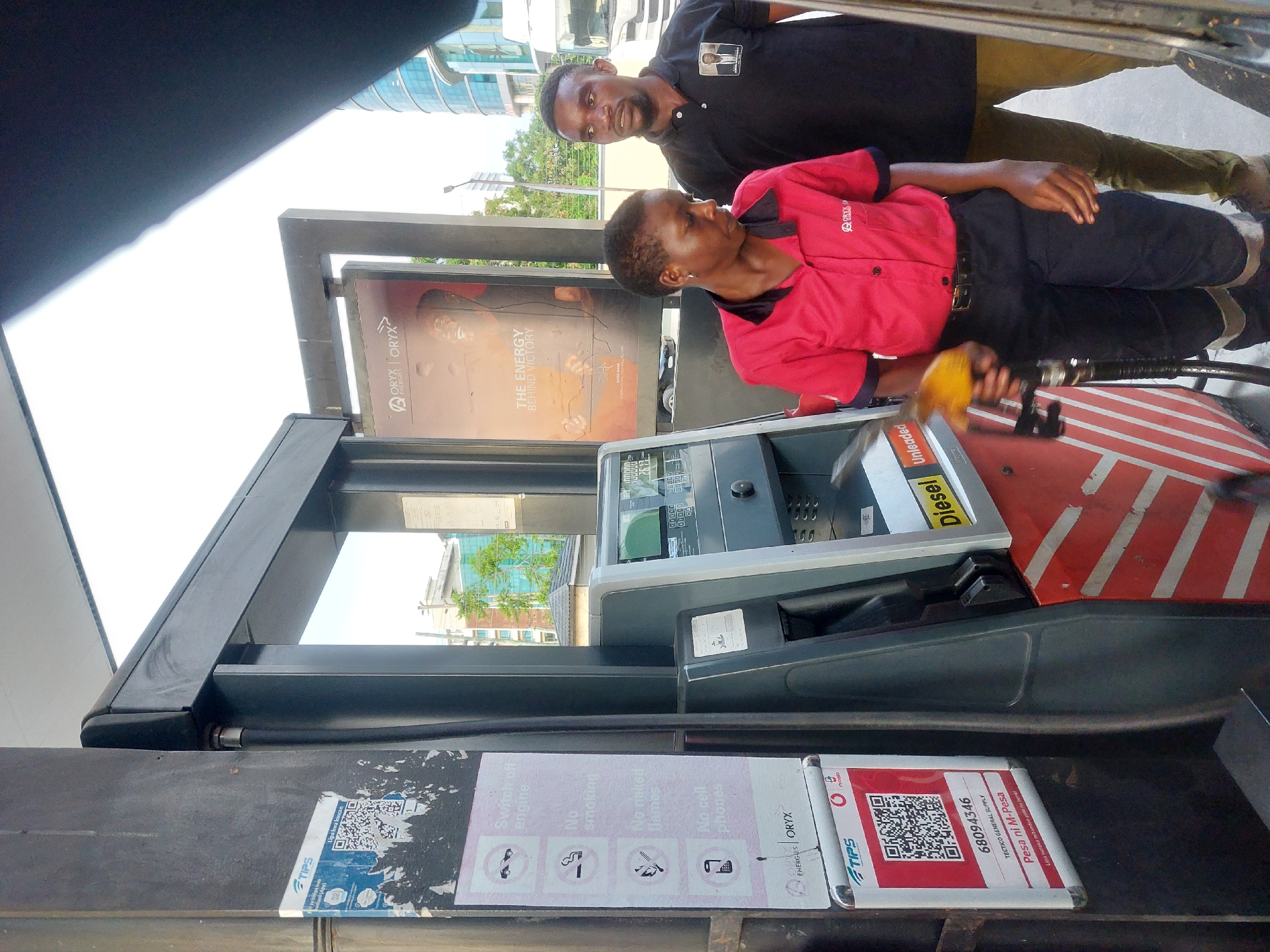 Here is a forensic description of the image:
**Scene:** A daytime scene at an Oryx Energies gas station in Dar es-Salaam, Tanzania.
**People:** Two African men. One man, is wearing a black polo shirt with an image and is standing next to the gas pump and behind the woman, and looking into the camera. The other man, is wearing a red Oryx Energies work uniform and black pants, is holding a yellow gas pump nozzle, appearing to be in the process of fueling a vehicle.
**Objects:** A gas pump with a digital display, signage indicating payment options, diesel unleaded and safety instructions, and Oryx Energies advertisements. There is a M-Pesa QR code on the gas pump.
**Actions:** The woman is holding a gas pump nozzle.
**Environment:** The scene appears to be a typical urban gas station environment, with building visible in the background. The overall lighting indicates a bright, sunny day.