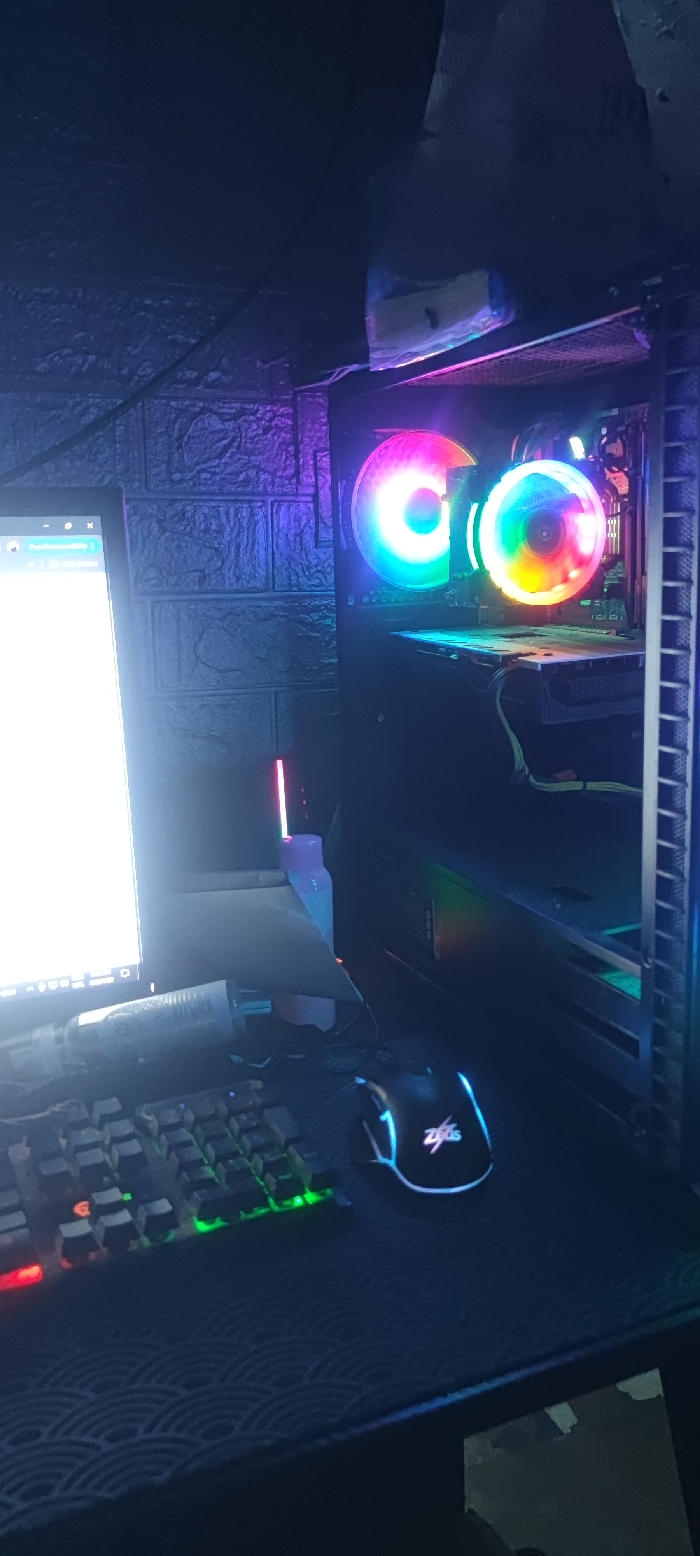 The image shows a computer desk with a computer, keyboard, mouse, and a tower. The tower is black and has RGB lighting. There is a laptop to the left and a mouse in the front. The desk is dark and the background is a dark brick wall. The location is in Dasmariñas, Philippines (latitude: 14.338, longitude: 120.947).