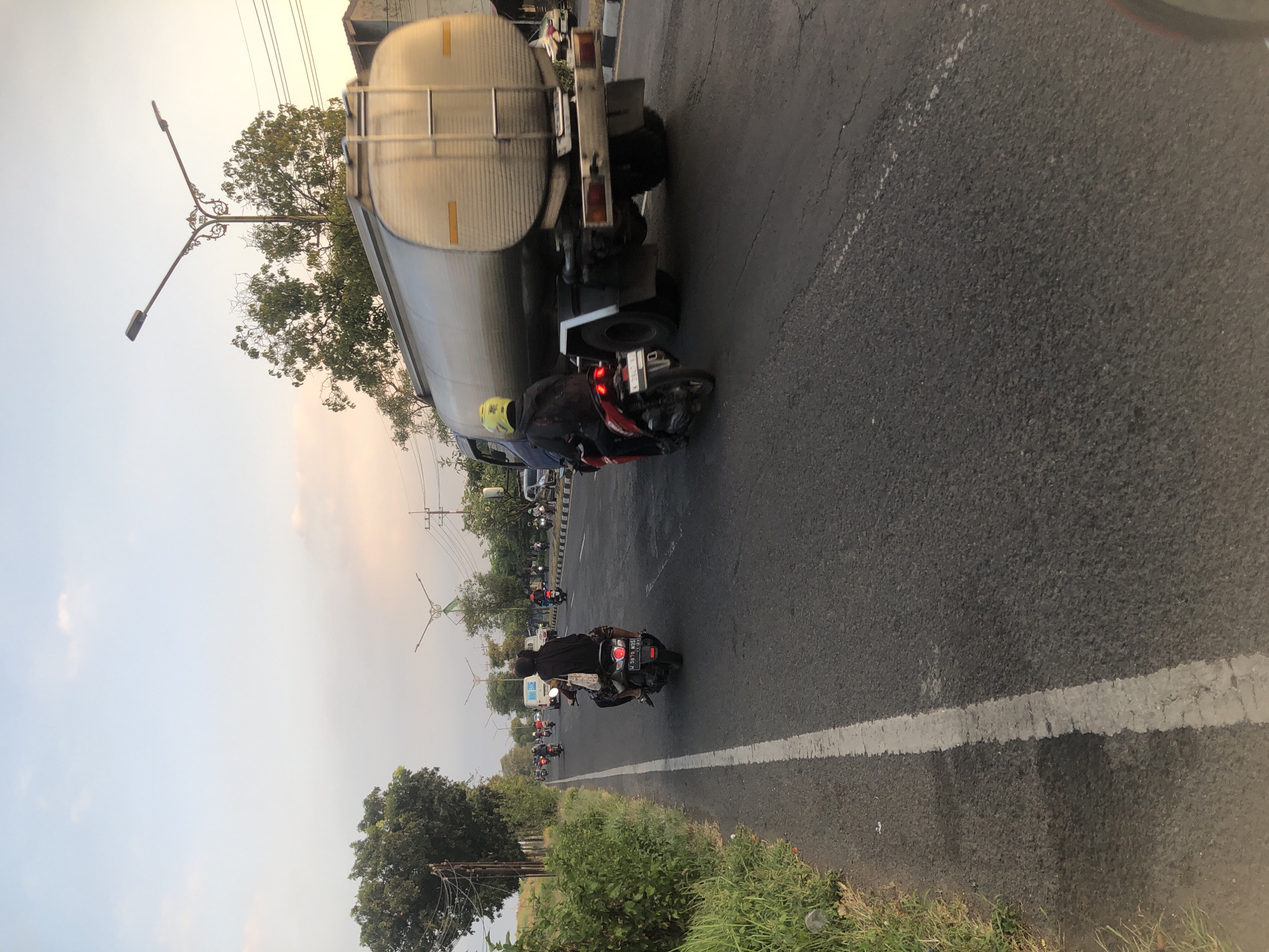 The image depicts a street scene in Siring, Indonesia, likely during the late afternoon or early evening due to the soft, warm light. The road is asphalt, marked with a white line on the left side, separating it from a grassy shoulder with green foliage. A large, silver tanker truck is the most prominent object, traveling on the right side of the road. In front of the truck, two motorcyclists are visible. The motorcyclist closer to the truck wears a yellow helmet and a dark jacket. The motorcyclist slightly further ahead wears a dark outfit and a black headscarf, and is carrying a passenger. Several other motorcycles are scattered further down the road, indicating moderate traffic. In the distance, trees and utility poles with streetlights can be seen, along with buildings on the right side of the road. The sky is a pale blue with hints of pink and orange near the horizon.